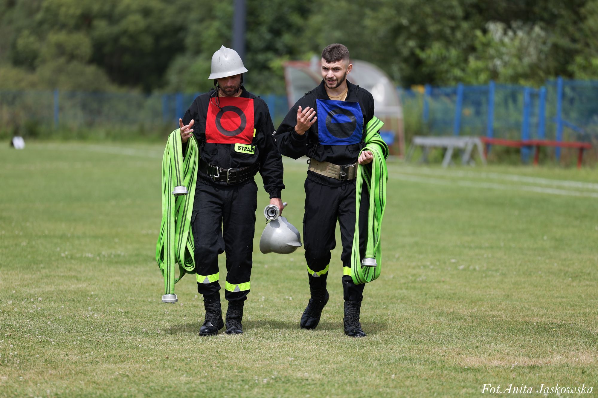 Dwóch mężczyzn w czarnych mundurach strażackich z odblaskowymi pasami, trzymający zielone zwinięte węże strażackie, jeden z nich w srebrnym hełmie.