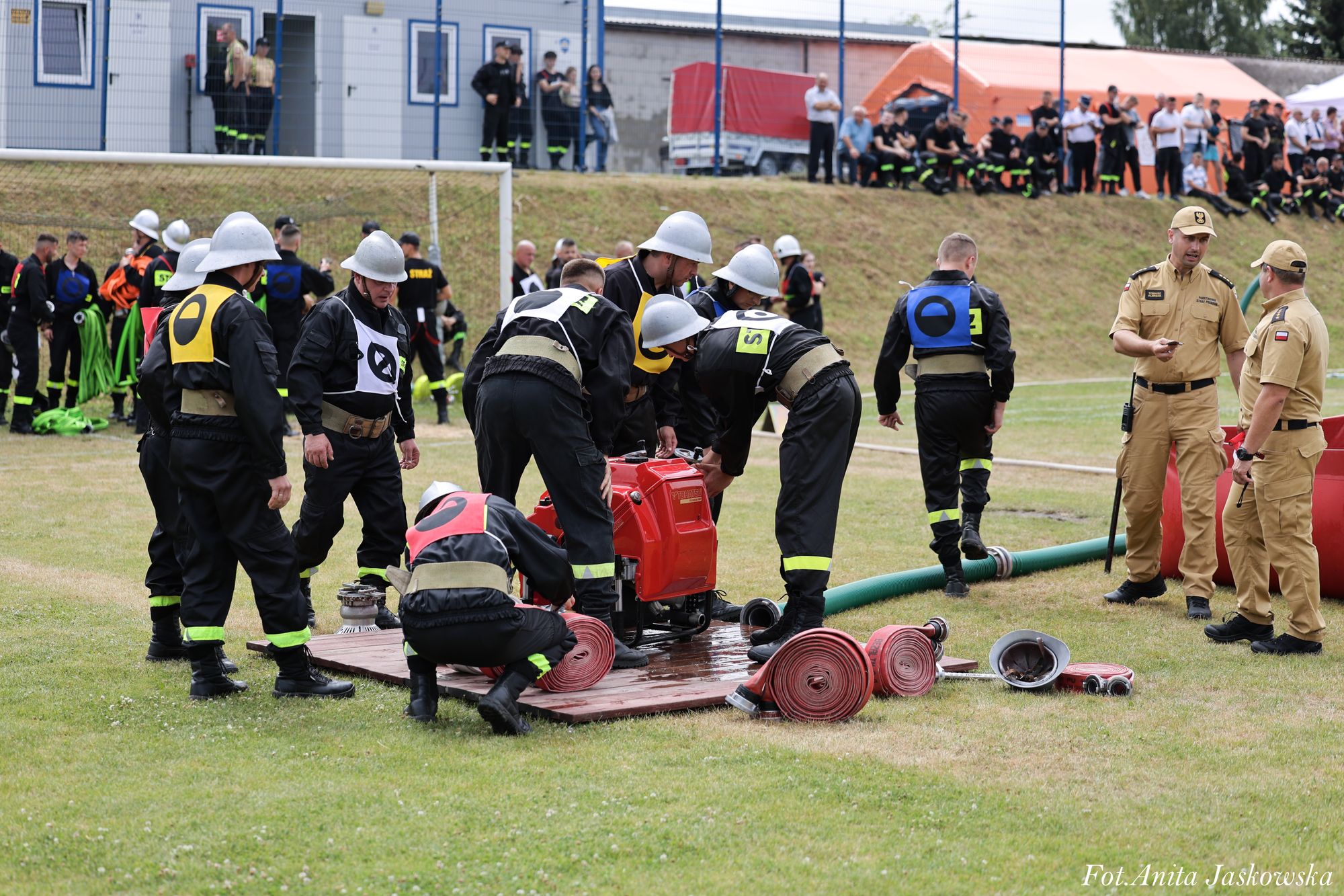Grupa strażaków w czarnych uniformach i białych hełmach przy rozłożonych czerwonych wężach i sprzęcie na trawie, w tle inni ludzie i budynki.