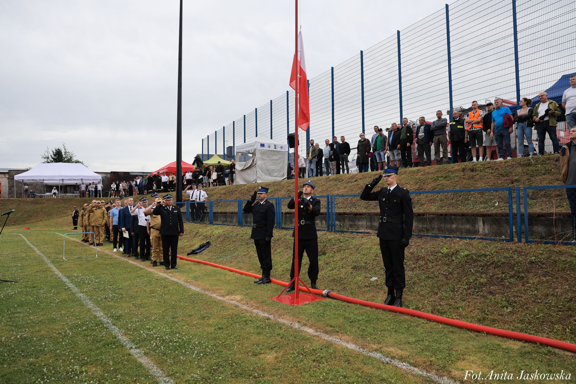 Trzech umundurowanych strażaków salutuje przy maszcie z biało-czerwoną flagą, za nimi w szeregu stoją ludzie w mundurach i cywilnych ubraniach na trawie, z tyłu tłum obserwatorów przy siatce.