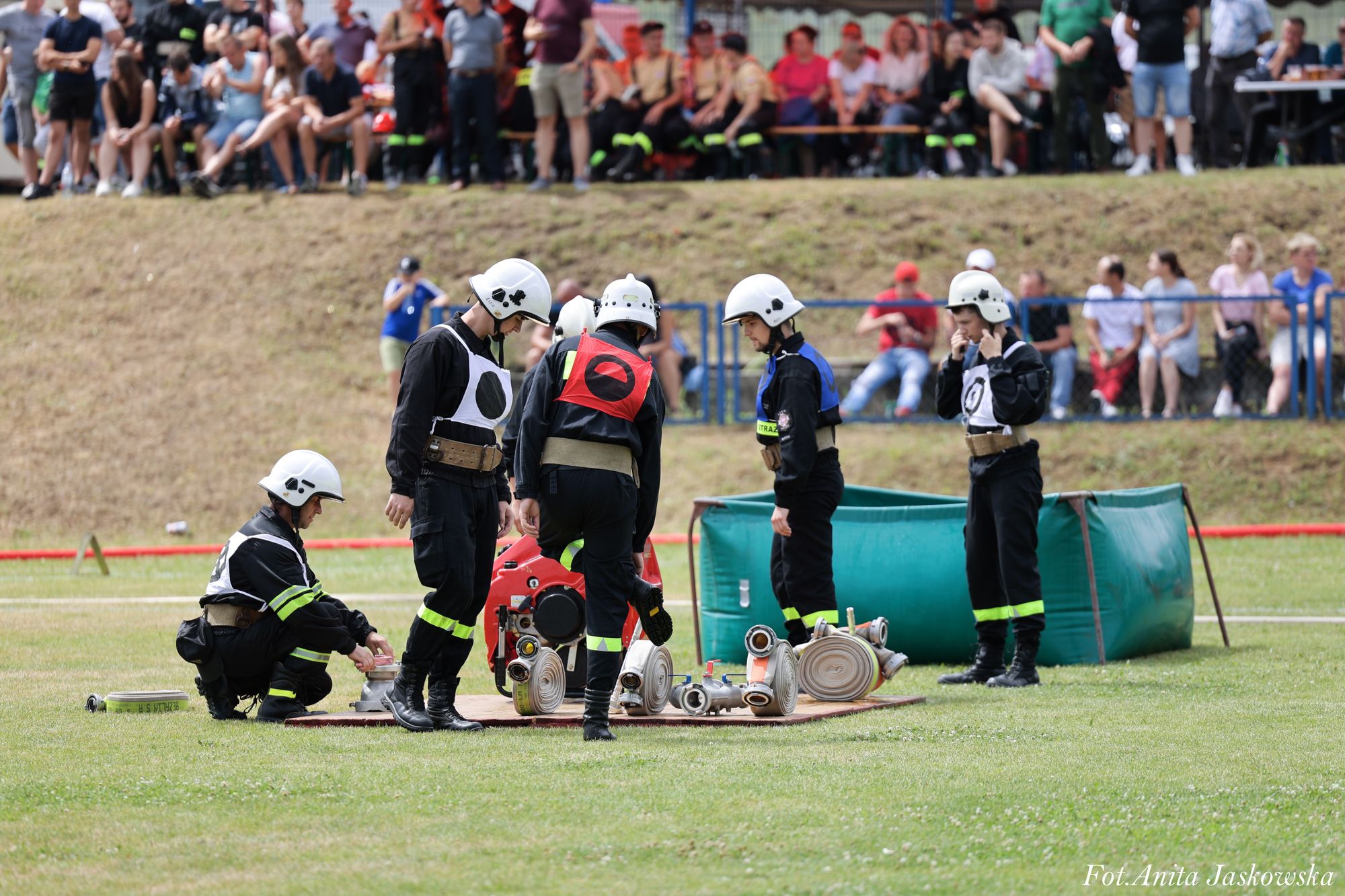 Pięciu mężczyzn w czarnych mundurach i białych hełmach stoi i klęczy na zielonej trawie obok złożonych zwiniętych węży strażackich, z tłumem ludzi w tle siedzących na ławkach.