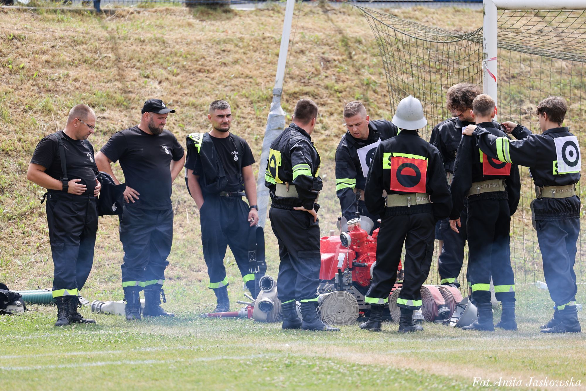 Grupa mężczyzn w czarnych mundurach z odblaskowymi paskami, stojących na trawie wokół czerwonej pompy pożarniczej i zwiniętych węży strażackich.
