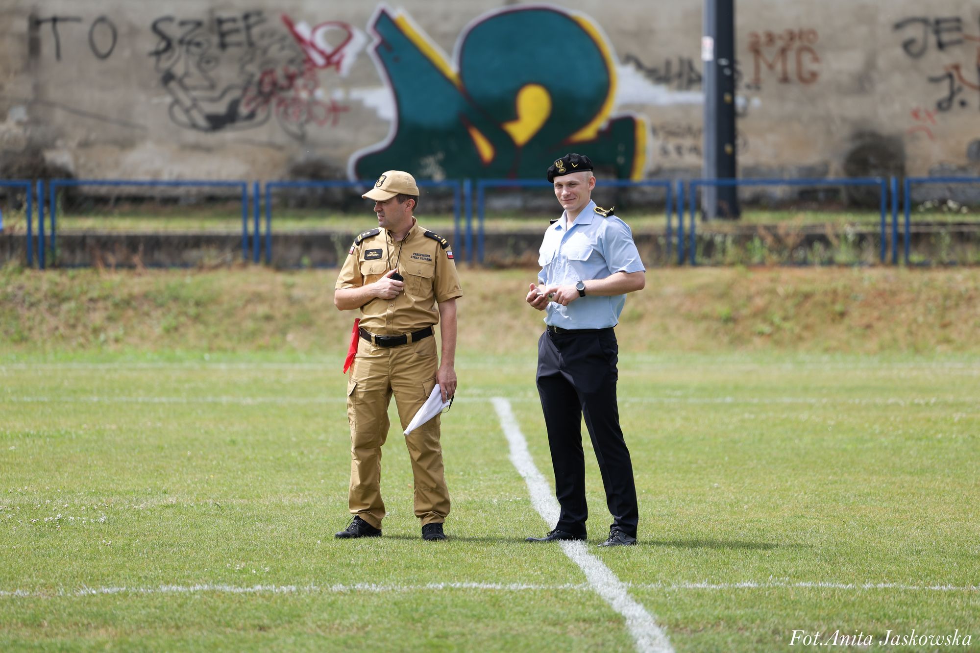 Dwóch mężczyzn w mundurach stoi na zielonym boisku sportowym, w tle mur z kolorowymi graffiti i niebieskie barierki.