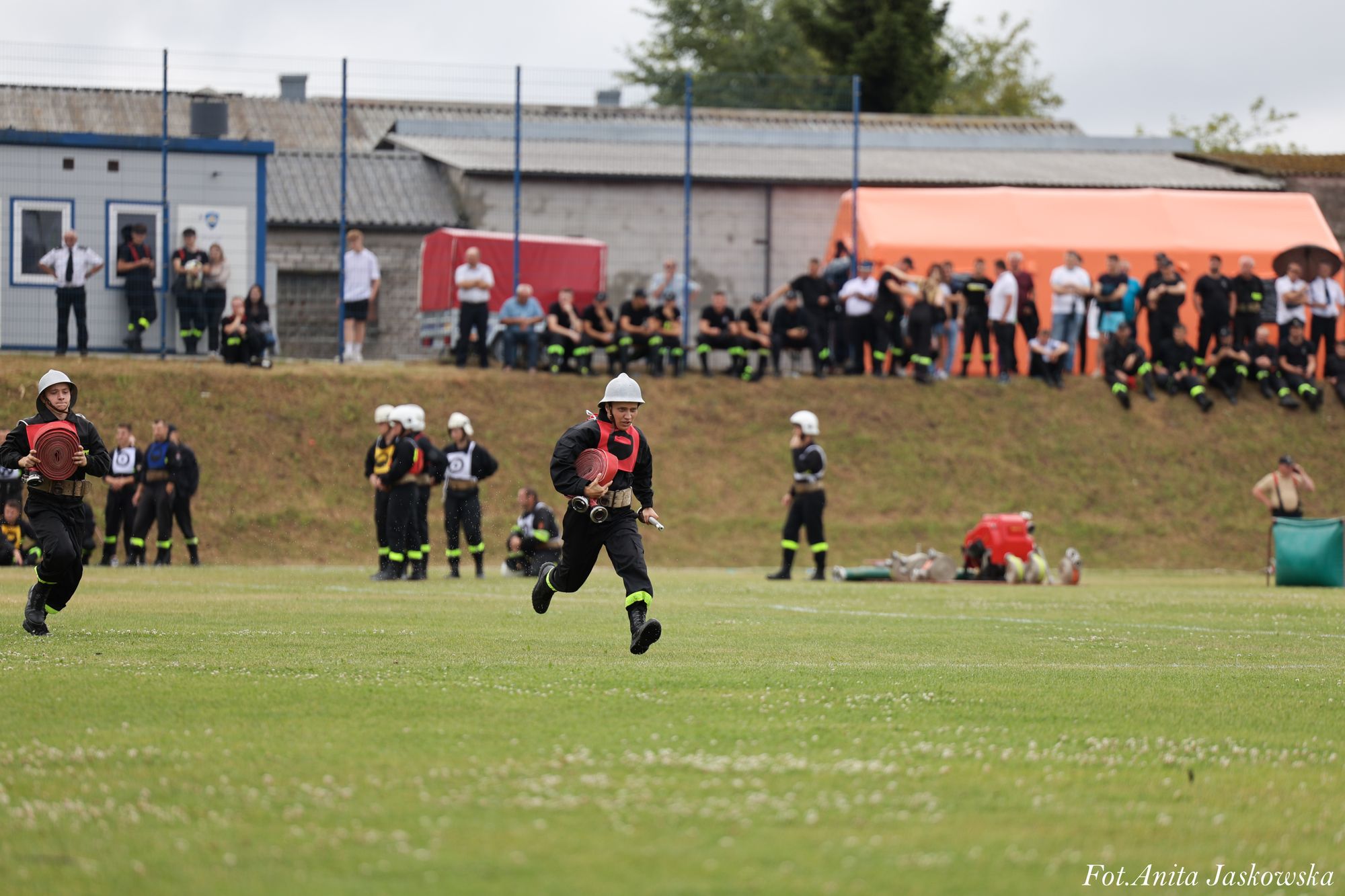 Dwóch strażaków w czarnych mundurach i białych hełmach biegnie po zielonym boisku, w tle grupa osób oraz pomarańczowy namiot i budynki.