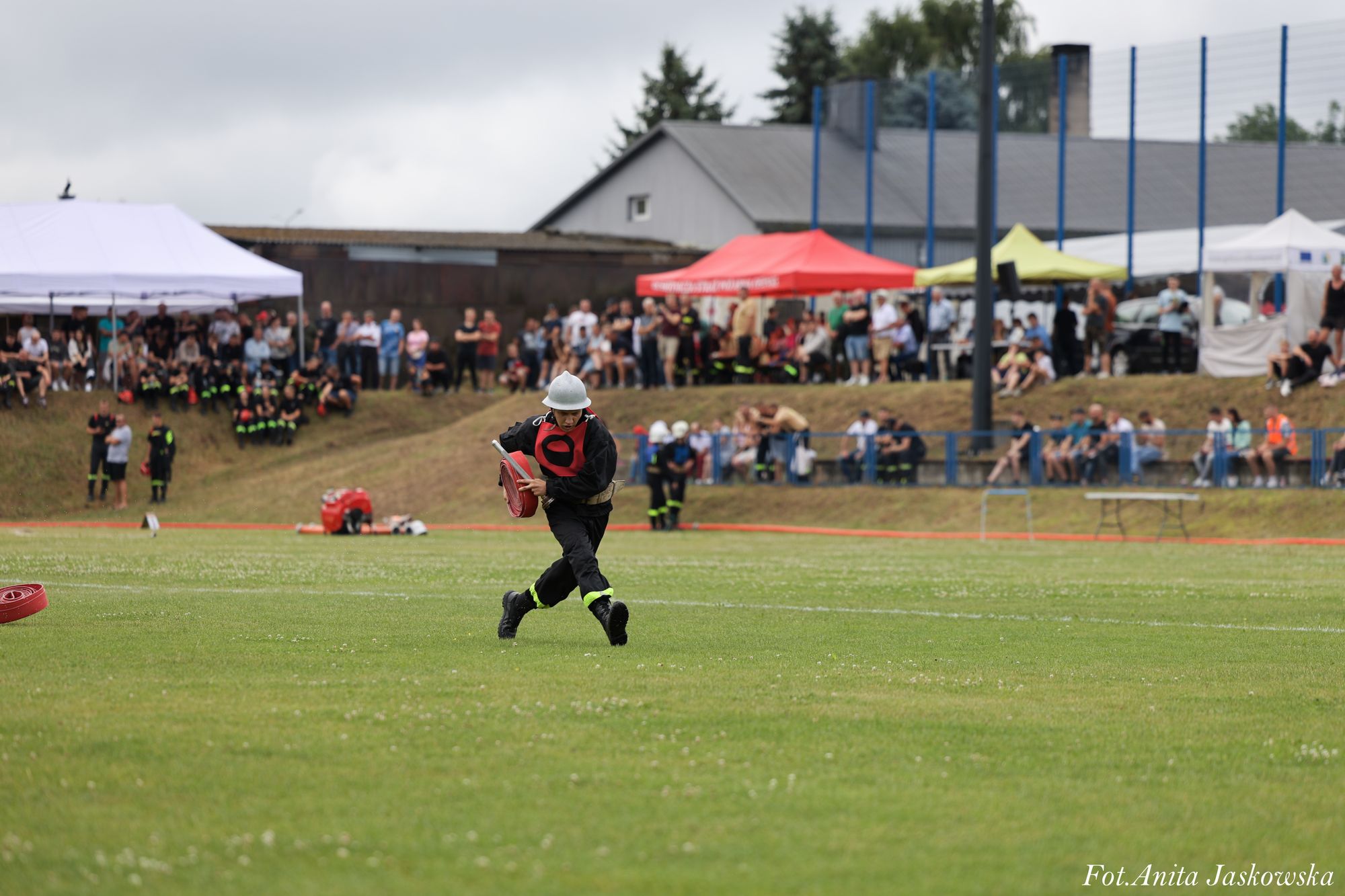 Młody strażak w czarnym ubraniu i srebrnym hełmie biegnie po zielonym boisku, trzymając zwinięty czerwony wąż strażacki, w tle tłum ludzi pod namiotami.