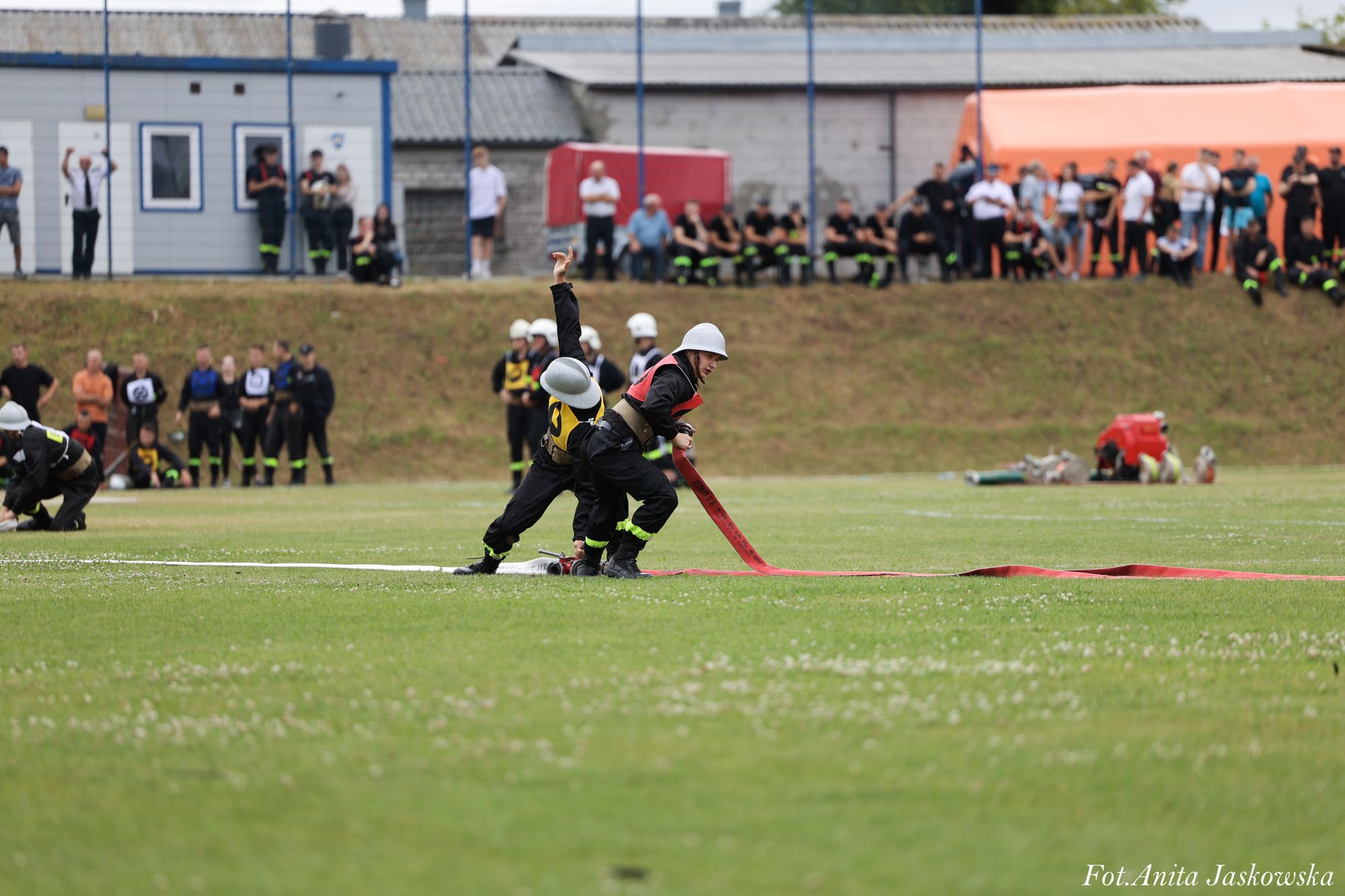 Dwóch strażaków w kaskach rozkłada czerwony wąż na zielonym boisku, w tle grupa osób obserwujących.