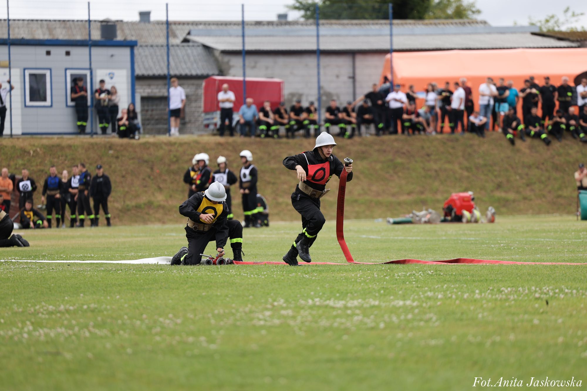 Dwóch strażaków w czarnych mundurach z żółtymi i czerwonymi oznaczeniami rozkłada czerwony wąż na zielonym boisku, w tle ludzie i budynki.