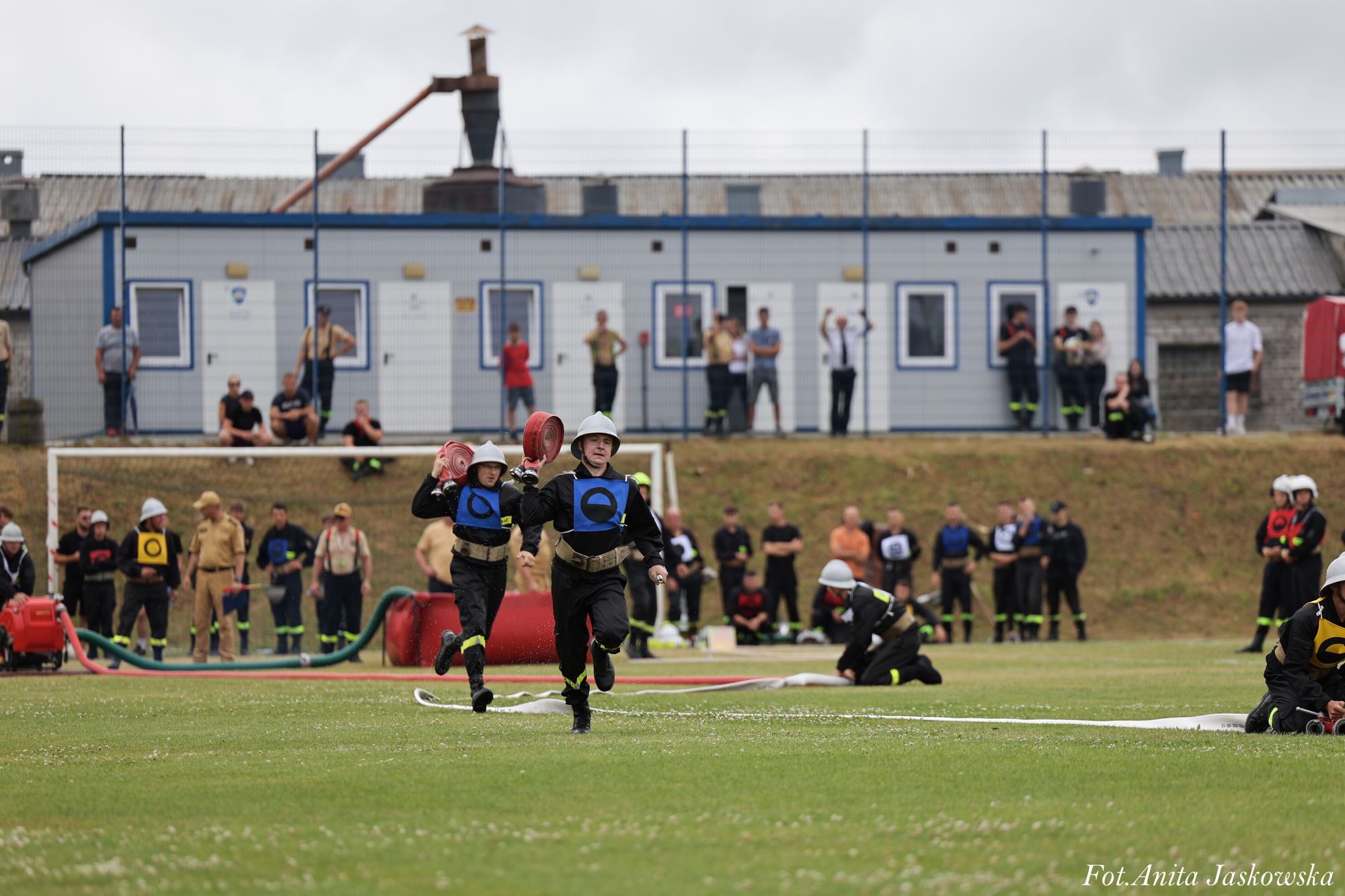 Dwóch strażaków w kaskach i niebieskich kamizelkach biegnie po zielonym boisku, trzymając czerwone zwinięte węże pożarnicze, w tle ludzie i budynki.