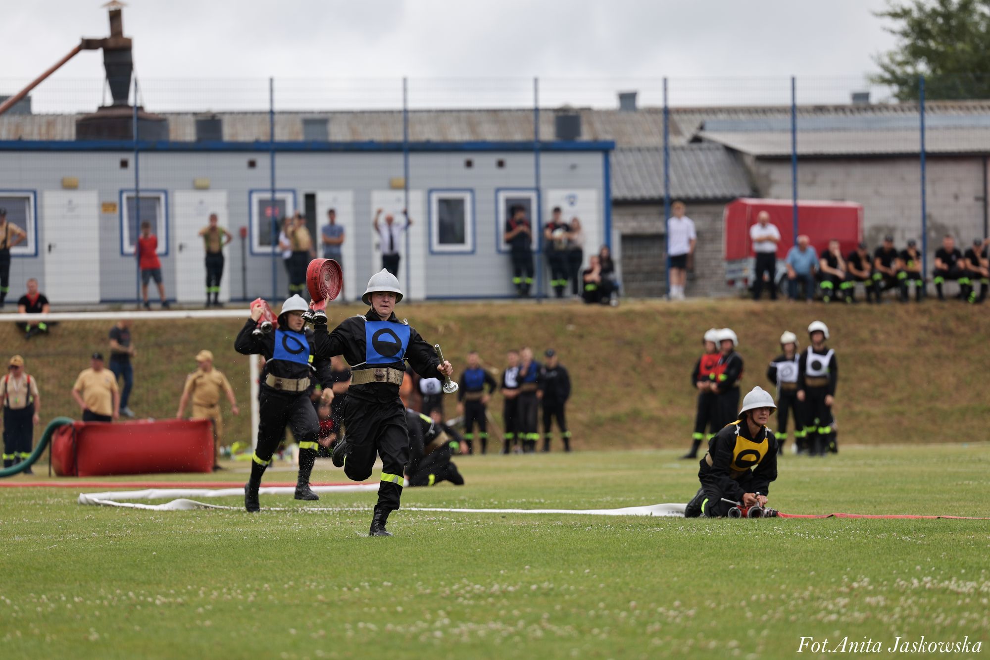 Strażacy w hełmach i czarnych ubraniach z kolorowymi oznaczeniami, biegnący i klęczący na zielonej trawie z rozwiniętymi wężami, w tle ludzie i budynki.