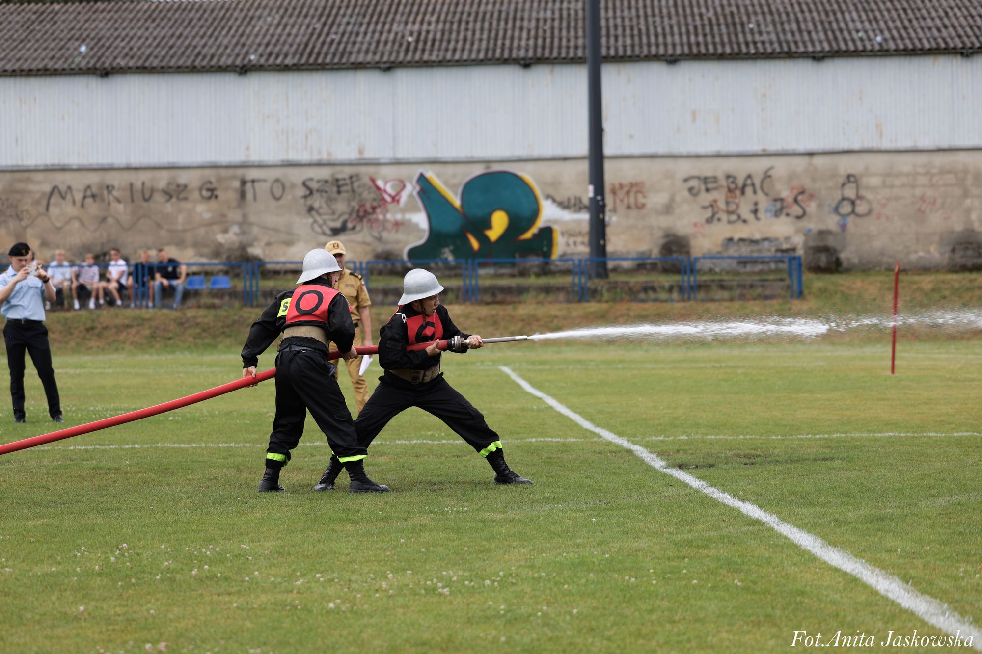 Dwóch strażaków w mundurach i hełmach trzyma czerwony wąż strażacki z wodą na zielonym boisku, w tle ludzie, ściana z graffiti i dach budynku.