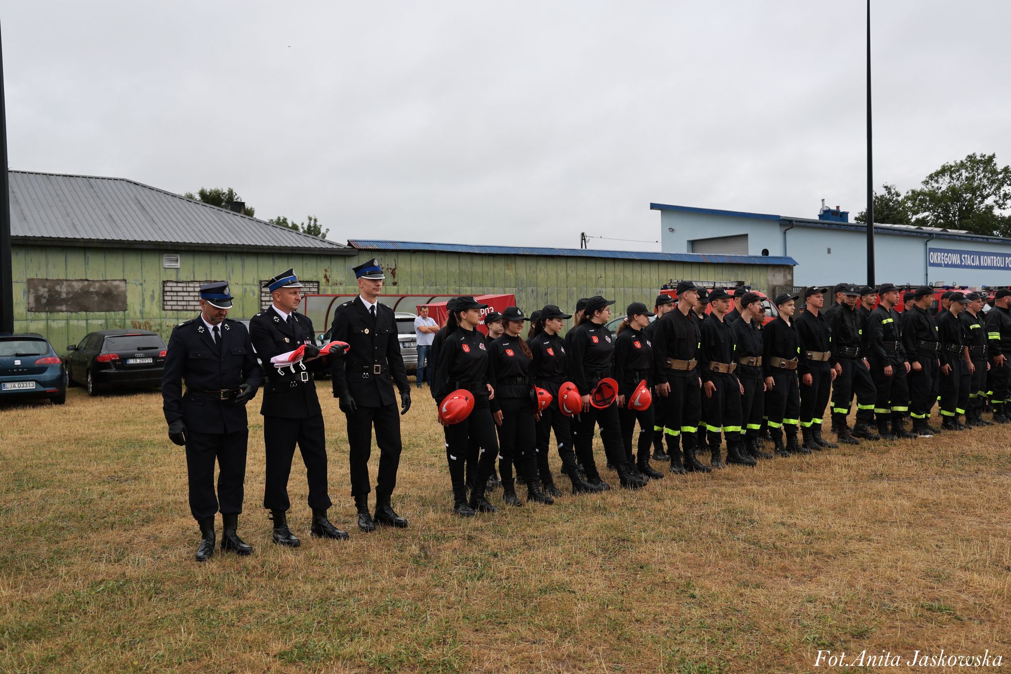 Grupa osób w mundurach strażackich ustawiona w szeregu na trawie, za nimi zielono-niebieskie budynki i zaparkowane samochody.