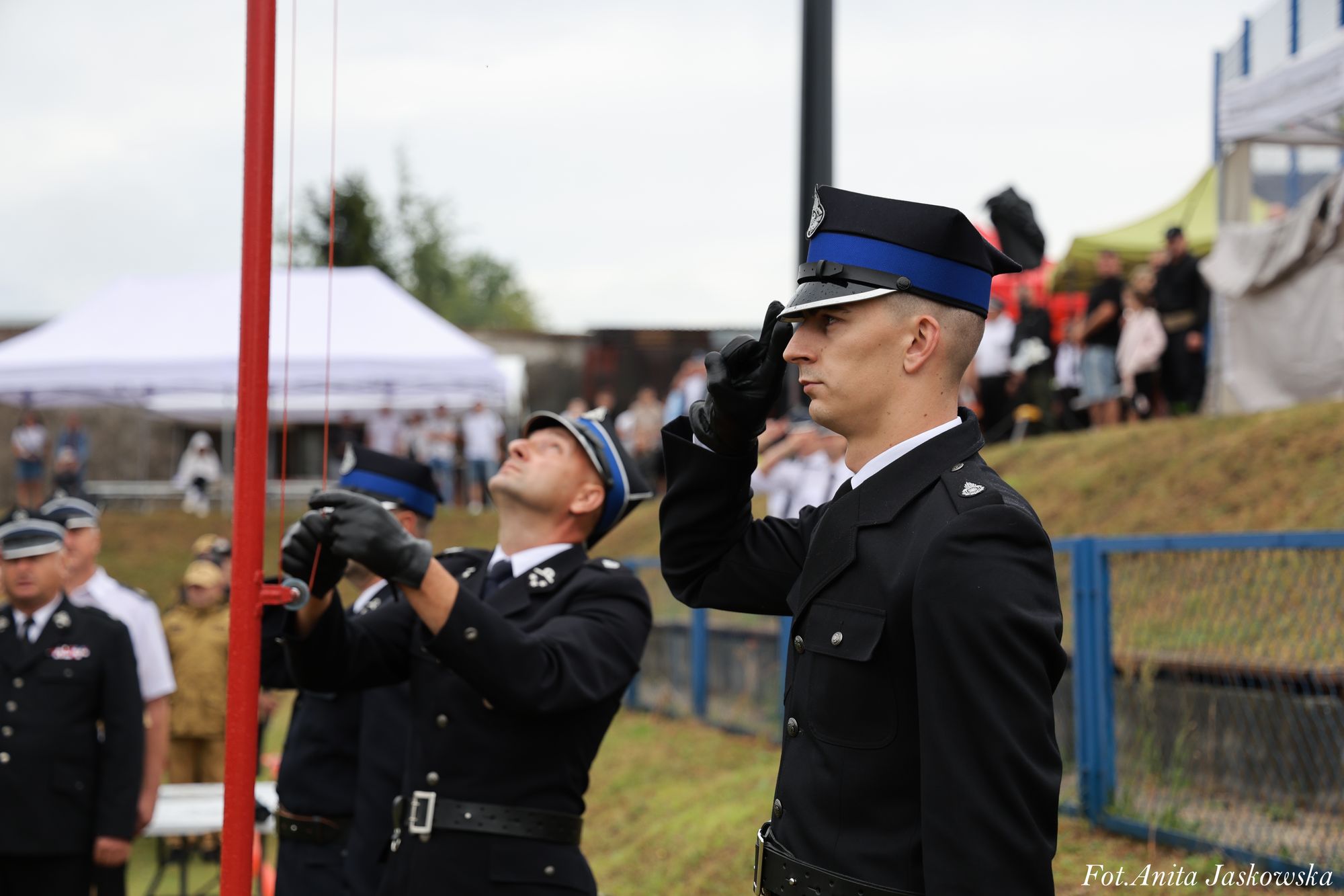 Mężczyźni w czarnych mundurach z niebieskimi lampasami, jeden salutuje, drugi podnosi flagę na maszcie, w tle rozmyta publiczność i namioty.