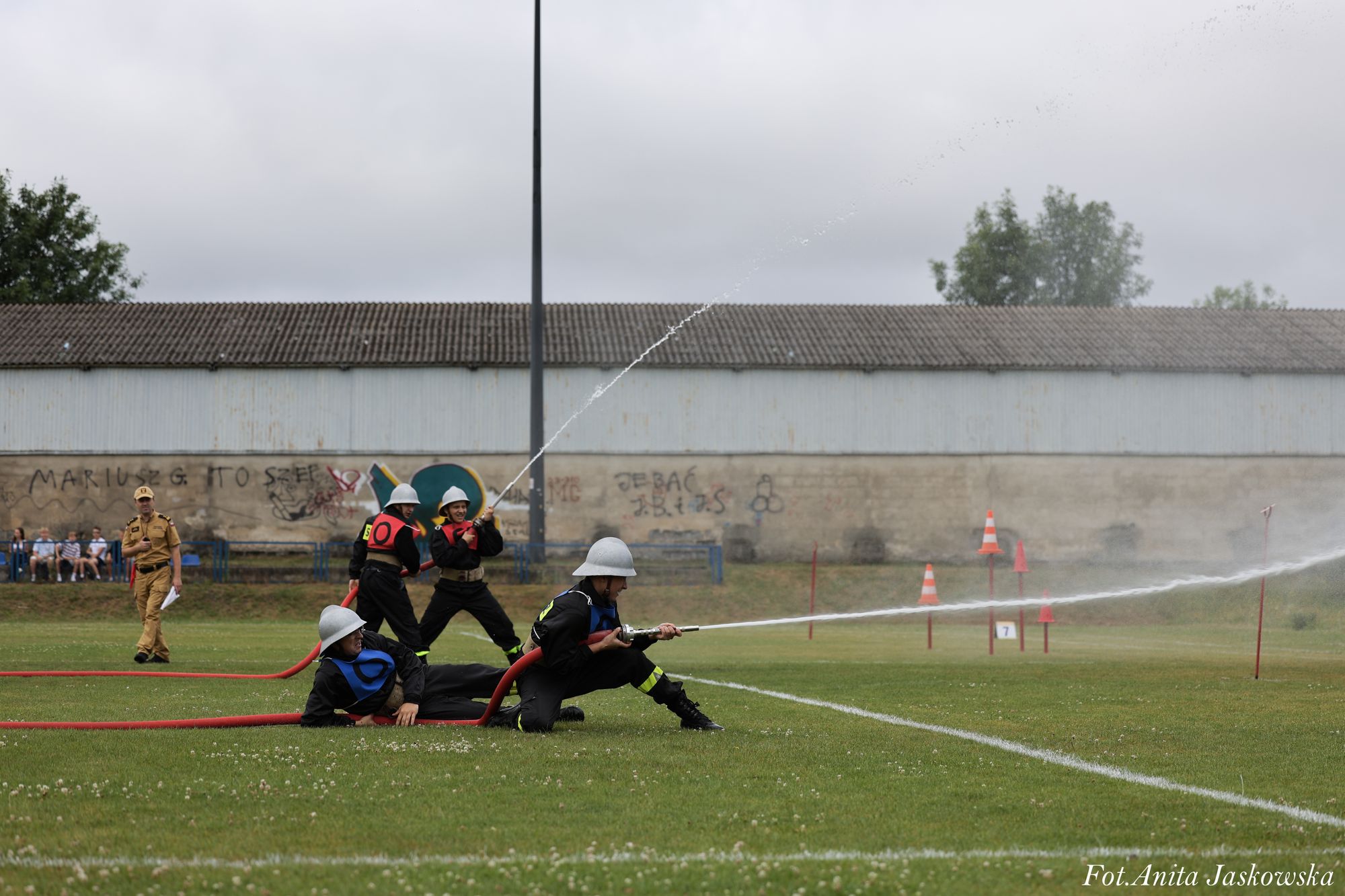 Cztery osoby w hełmach i mundurach strażackich na zielonej trawie, dwie z nich trzymają i kierują wodę z węży strażackich, w tle mur z graffiti i stojący mężczyzna w brązowym mundurze.