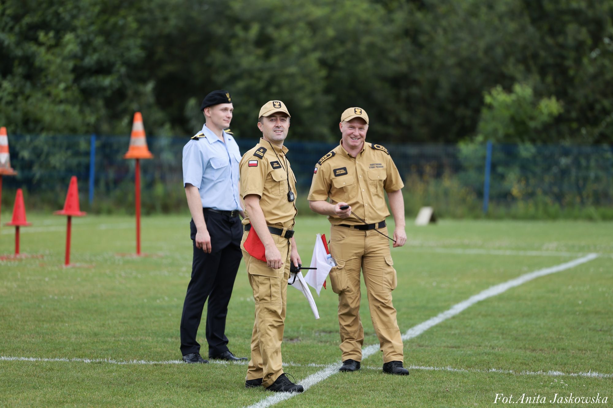 Trzech mężczyzn w mundurach na zielonym boisku, dwóch w beżowych mundurach z czapkami, jeden w jasnoniebieskiej koszuli i czarnym berecie, w tle pomarańczowe pachołki i zieleń.