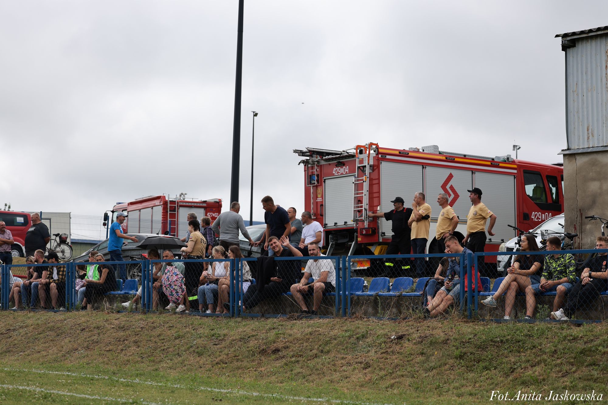 Ludzie siedzący na niebieskich ławkach za niebieskim metalowym ogrodzeniem, w tle dwa czerwone wozy strażackie i szara budowla, pochmurne niebo.