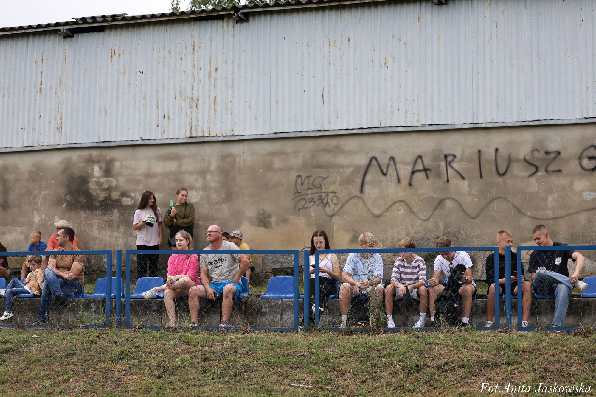 Grupa osób siedzących na niebieskich krzesłach za niebieską metalową barierką na tle szarej, częściowo zniszczonej ściany z czarnym graffiti.
