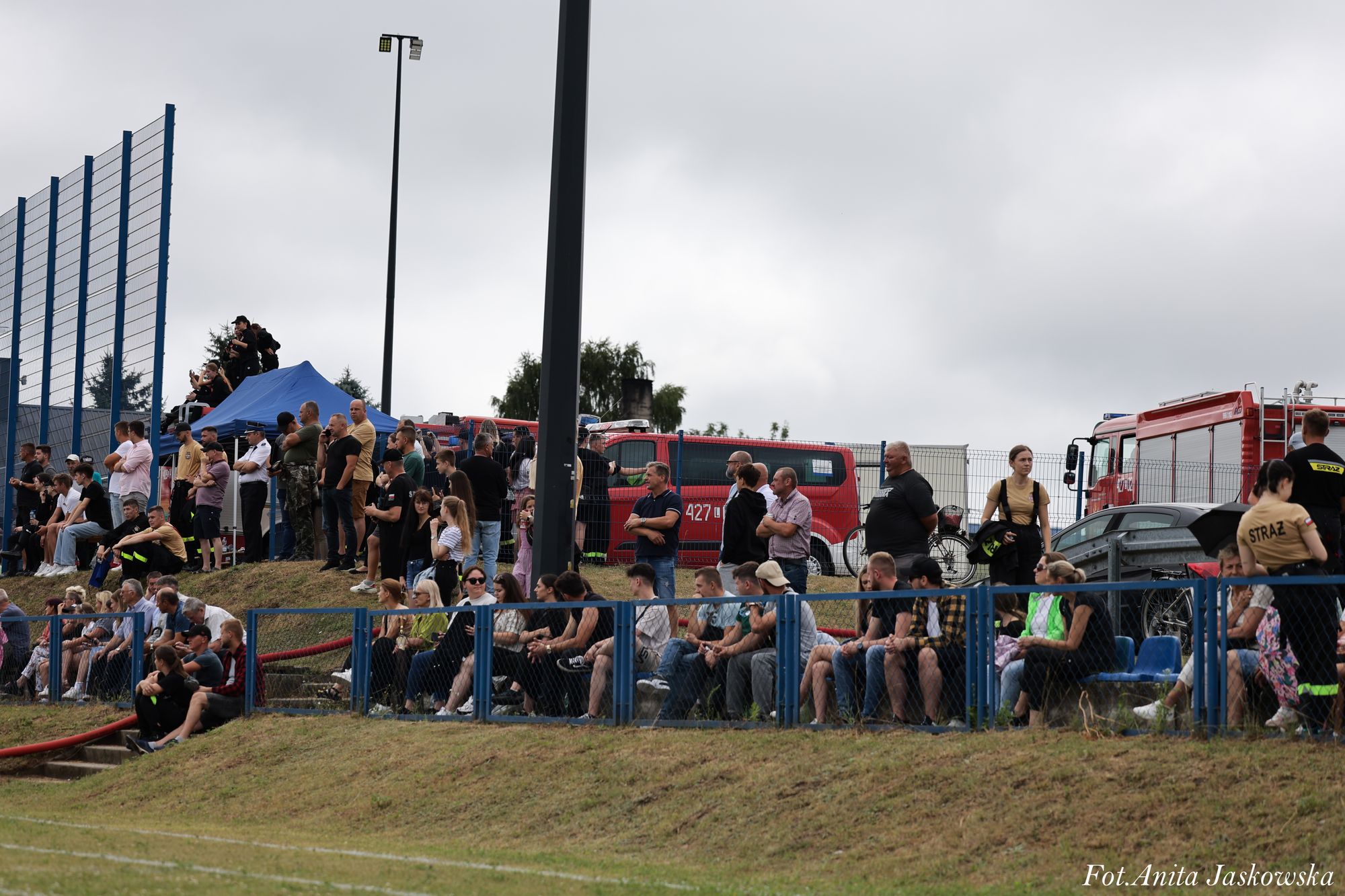 Ludzie siedzący i stojący na trawiastym wzniesieniu, niebieskie barierki i płot, czerwone wozy strażackie, pochmurne niebo.