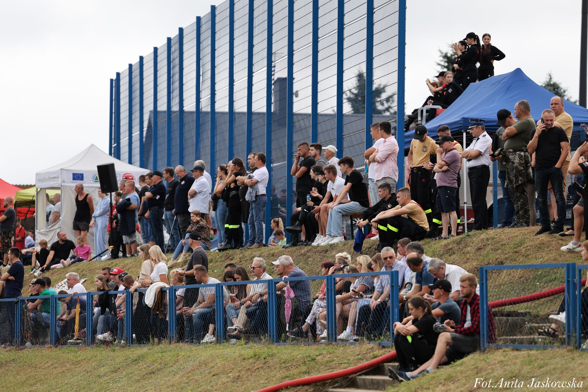 Grupa ludzi siedzi i stoi na trawiastej skarpie przy niebieskim ogrodzeniu, w tle namioty i wysoka siatka z niebieskimi słupkami.