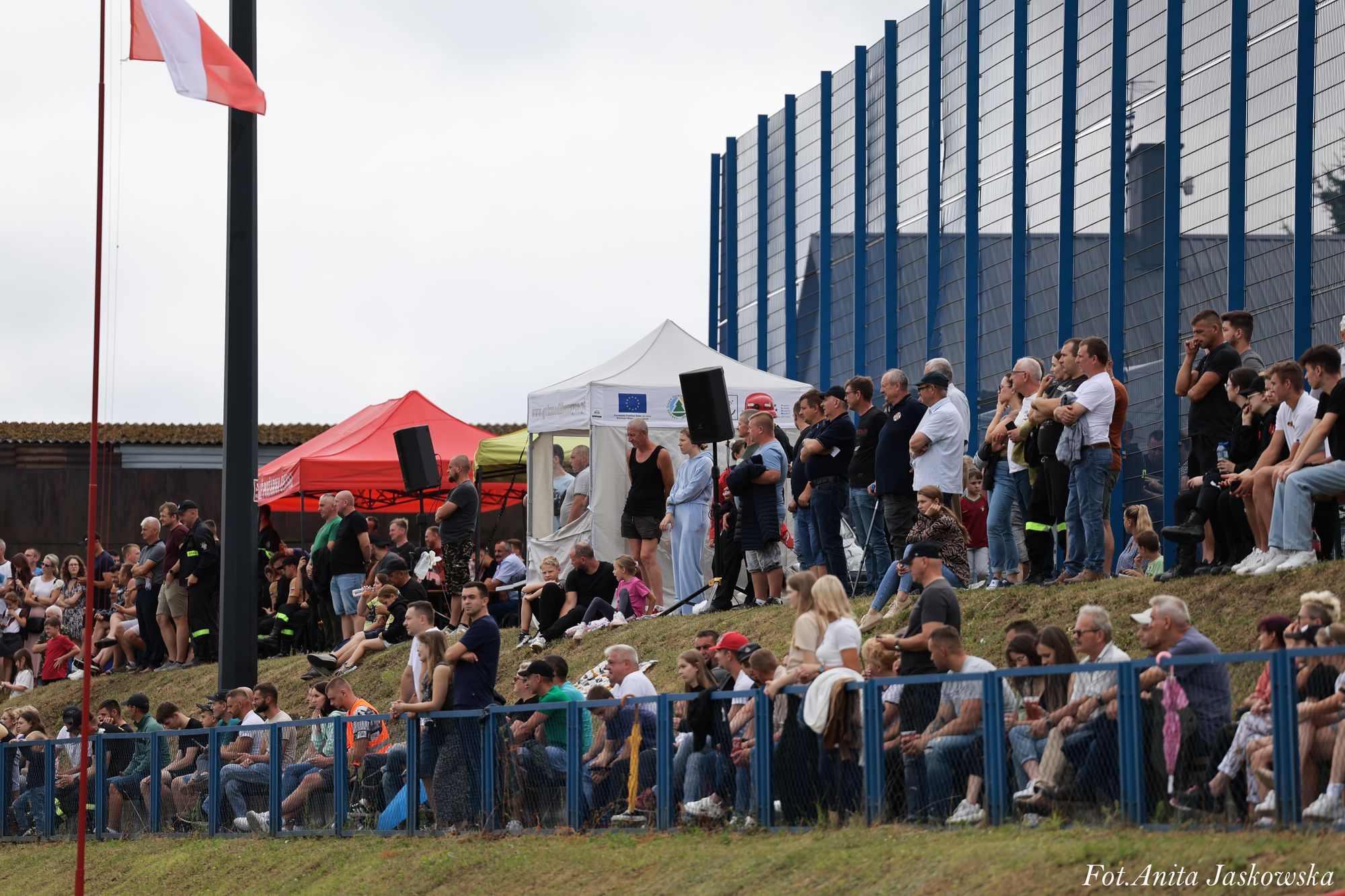 Ludzie stojący i siedzący na trawiastym wzniesieniu przy niebieskim ogrodzeniu, biało-czerwona flaga na maszcie, czerwone i białe namioty, szklana ściana w tle.