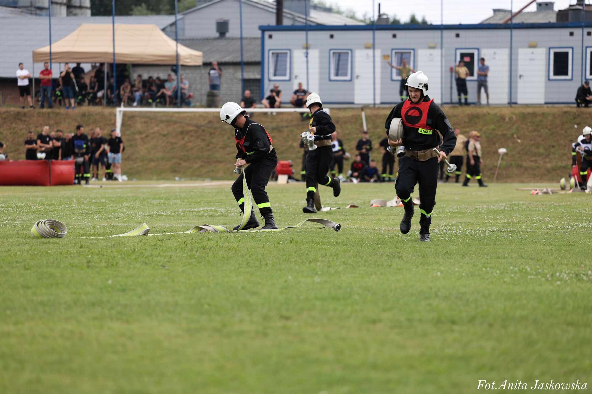 Strażacy w czarnych mundurach z białymi hełmami rozkładają zwinięte węże na zielonej trawie, w tle ludzie i namiot na boisku sportowym.