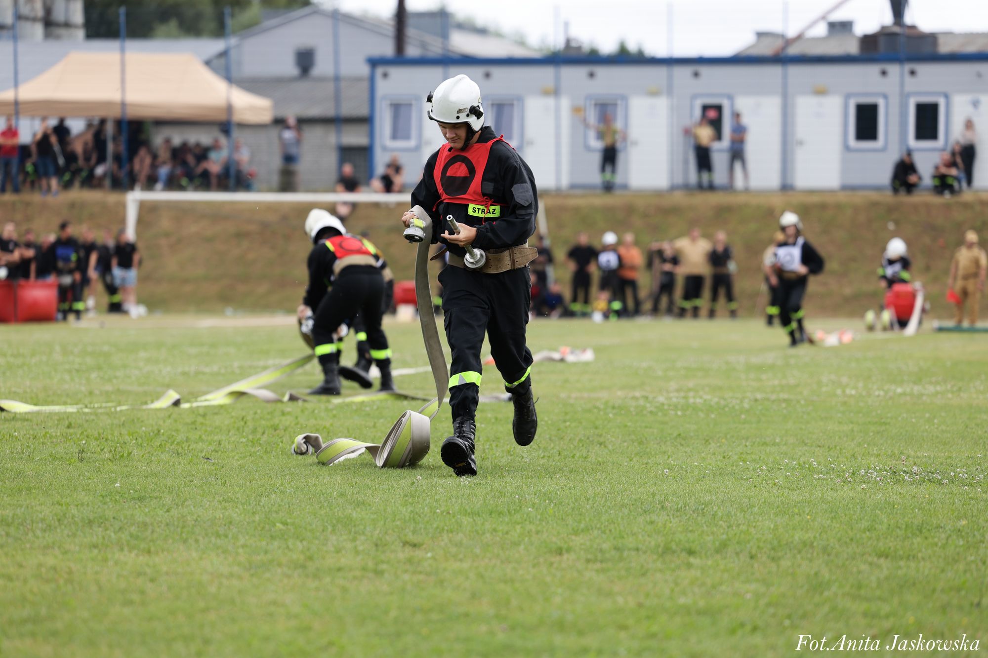 Strażak w czarnym ubraniu z czerwonym oznaczeniem i białym hełmem biegnie po trawie, trzymając rozwinięty wąż strażacki, w tle inne osoby i budynki.