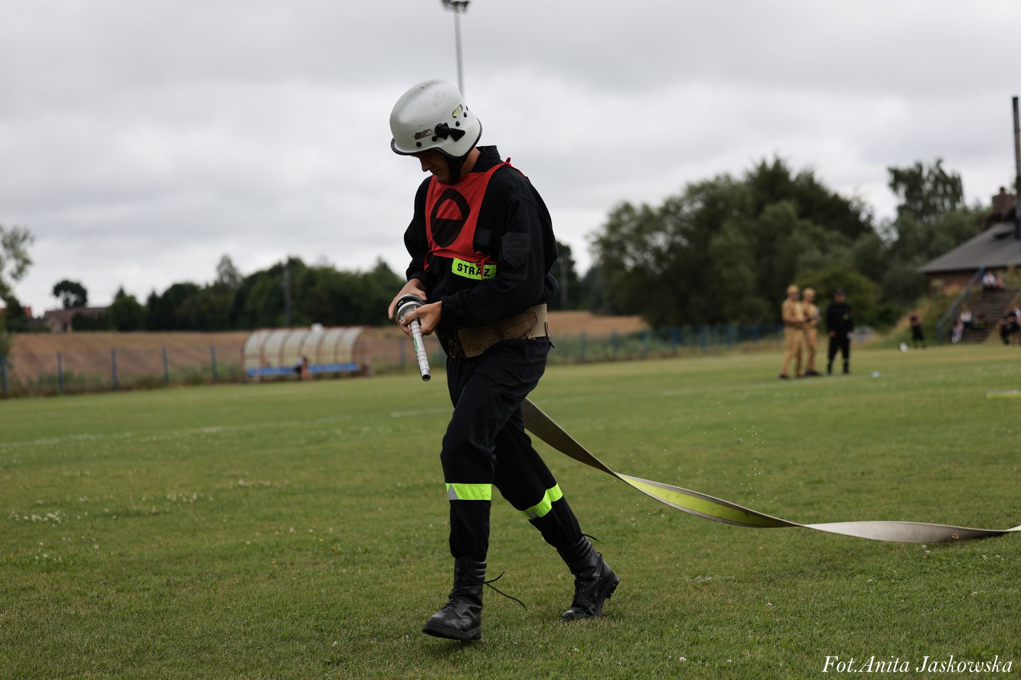 Strażak w czarnym uniformie z odblaskowymi paskami i białym hełmem trzyma wąż strażacki na zielonym boisku, w tle drzewa i ludzie.