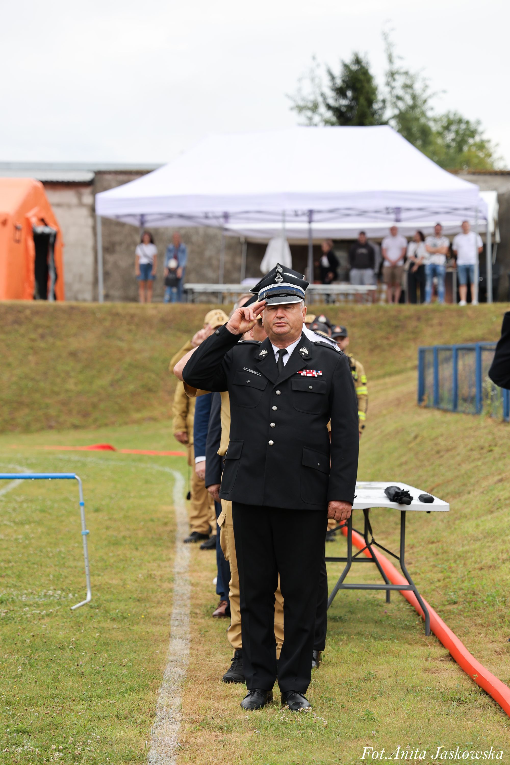 Mężczyzna w czarnym mundurze z czapką na głowie stoi na linii białej na trawiastym boisku, za nim szeregiem ustawieni inni mężczyźni w mundurach. W tle biały namiot i ludzie.