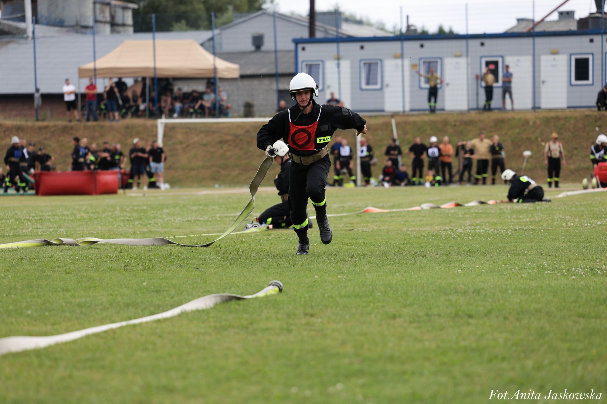Strażak w czarnym ubraniu i białym hełmie biegnie po zielonym trawniku, trzymając rozciągnięty wąż strażacki, w tle ludzie i budynki.