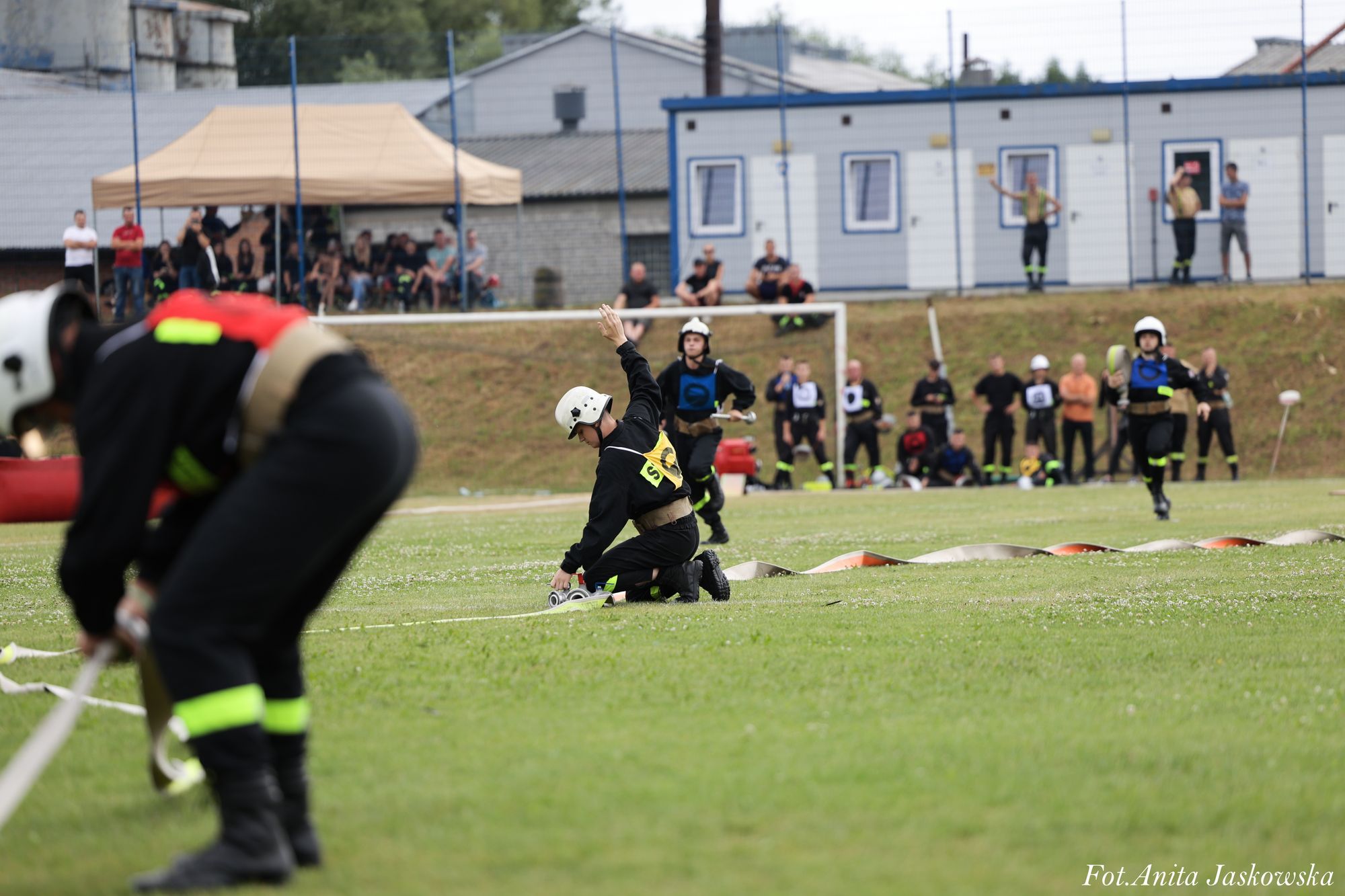 Strażacy w czarnych mundurach z odblaskowymi elementami i białych hełmach przygotowują sprzęt na zielonym boisku, w tle ludzie pod beżowym namiotem i budynki.