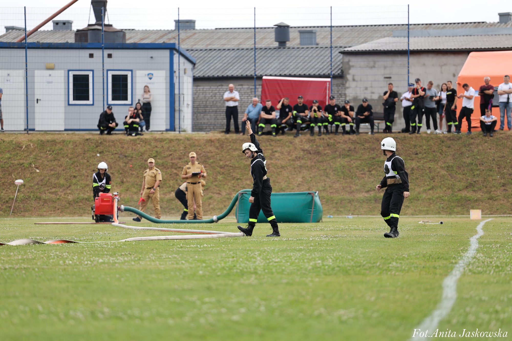 Trzech strażaków w czarnych uniformach i białych hełmach na zielonym boisku, w tle grupa ludzi i budynki, na pierwszym planie wąż strażacki i sprzęt.