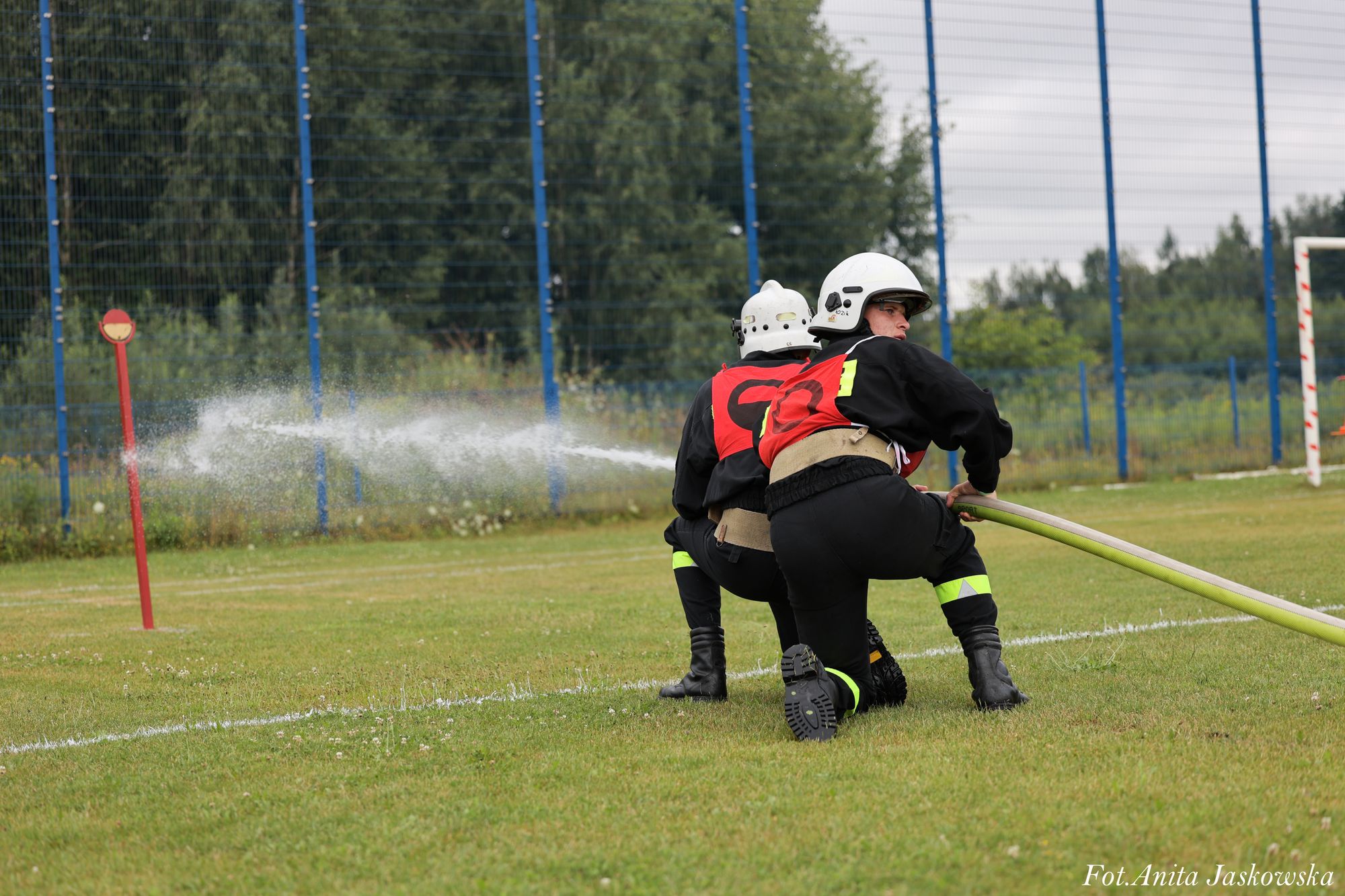 Dwóch strażaków w hełmach i czerwonych kamizelkach z wężem gaszącym wodą na zielonym boisku sportowym.