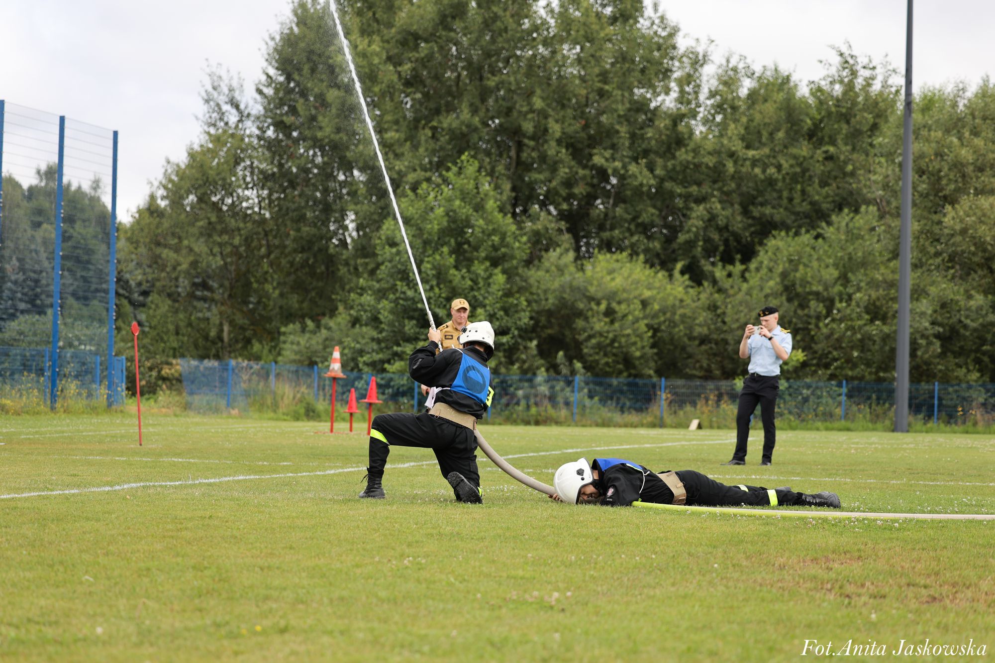 Dwóch strażaków w kaskach na zielonym boisku, jeden klęczy i trzyma strumień wody, drugi leży na trawie z rurą strażacką, w tle dwie osoby obserwujące.