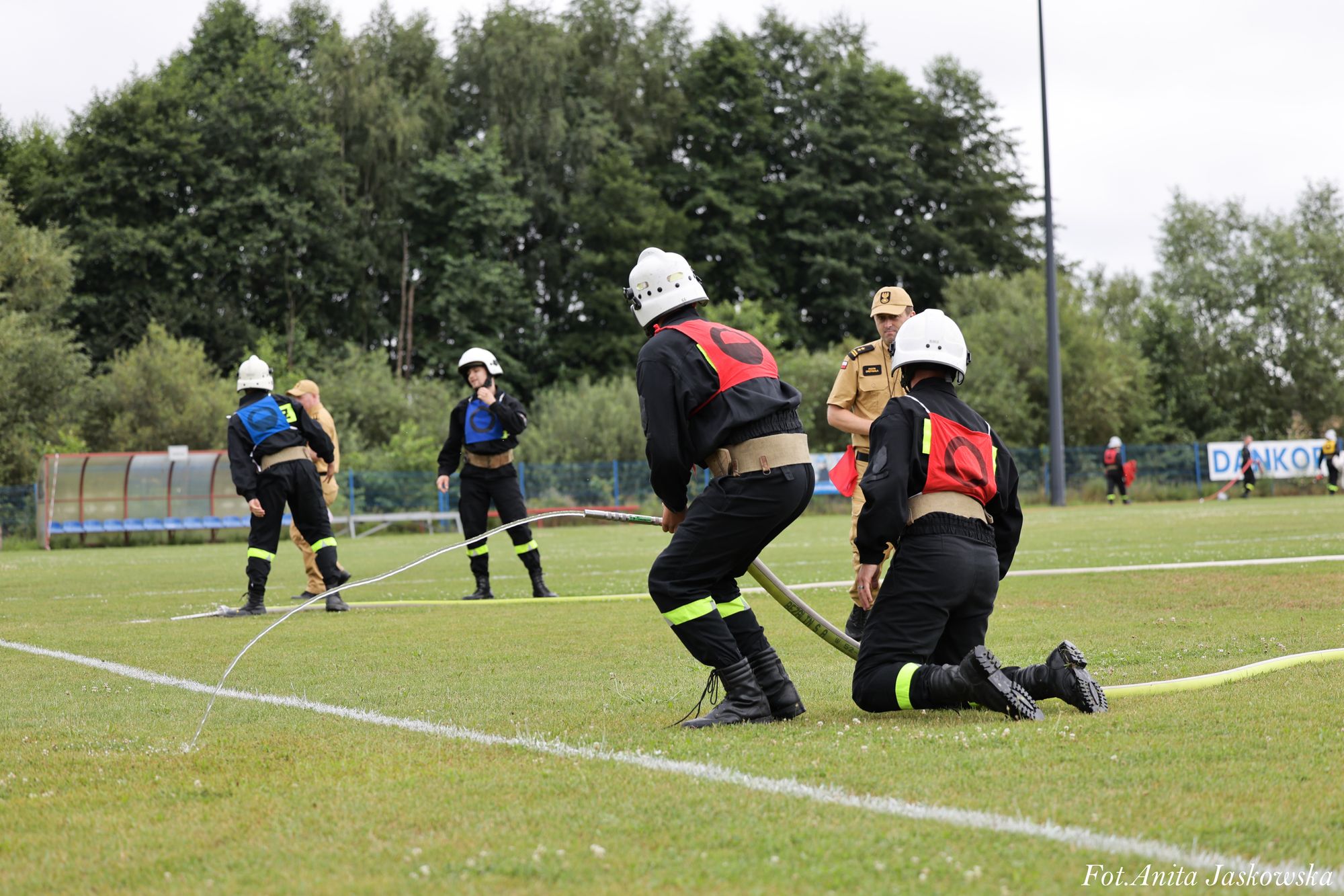 Cztery osoby w mundurach strażackich z kaskami na zielonym boisku, dwie trzymające prąd wody z węża, w tle las i sędzia w beżowym ubraniu.