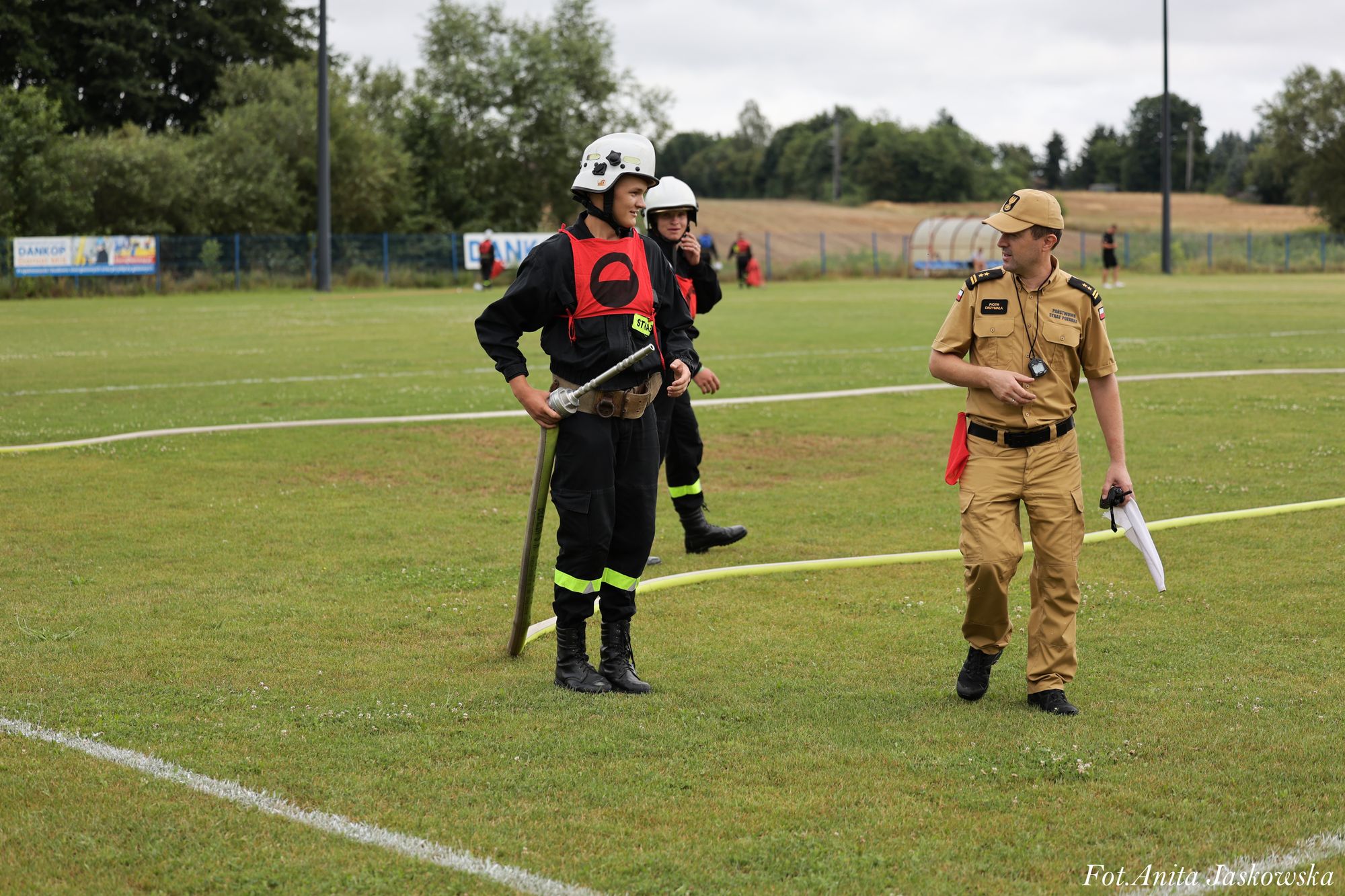 Dwóch mężczyzn w czarnych mundurach strażackich z czerwonymi oznaczeniami i hełmami stoi na trawie, obok nich mężczyzna w beżowym mundurze z czapką i akcesoriami.