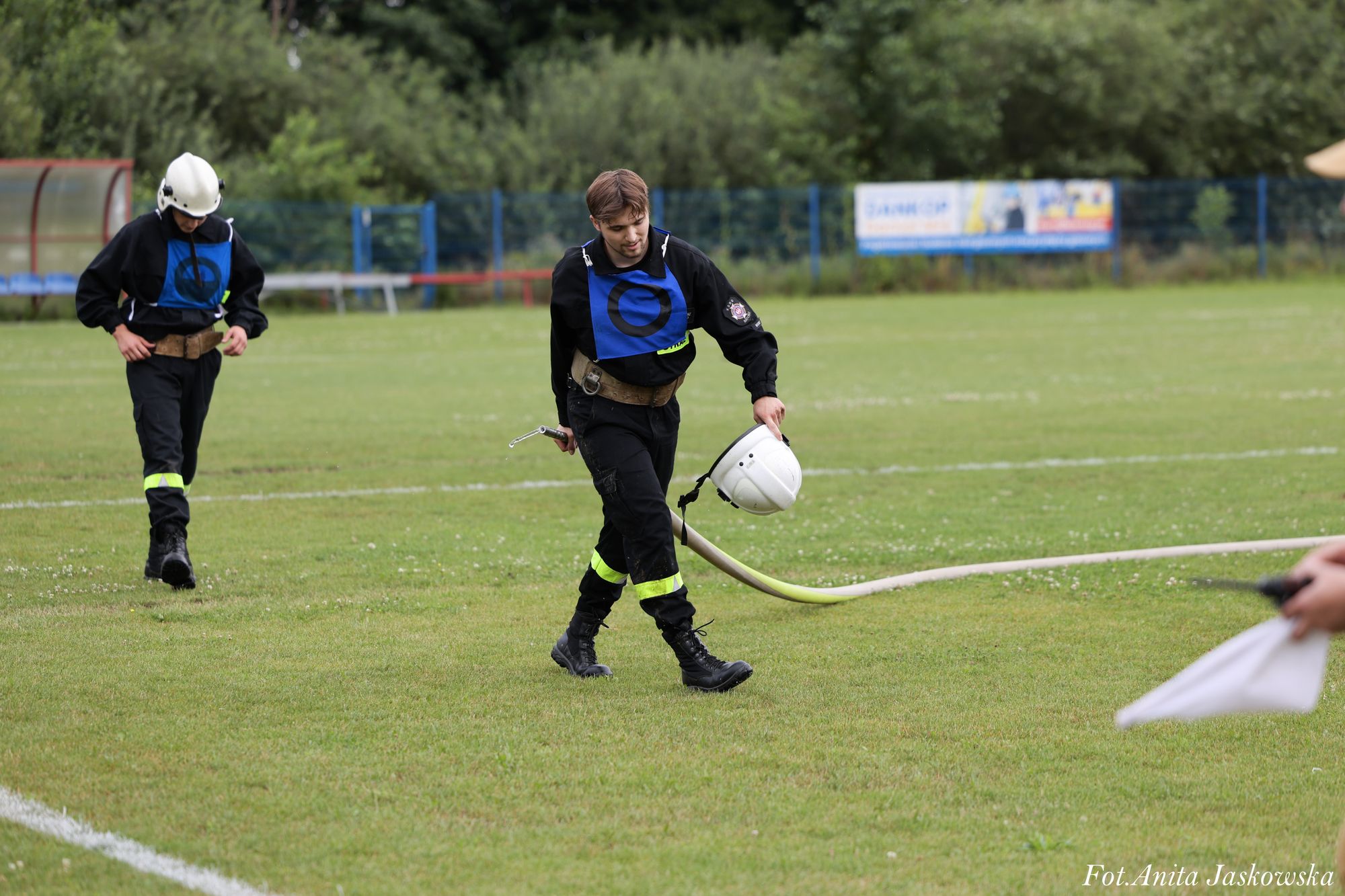 Dwóch mężczyzn w czarnych ubraniach z niebieskimi oznaczeniami, jeden trzyma biały hełm i wąż strażacki na zielonej trawie boiska sportowego.