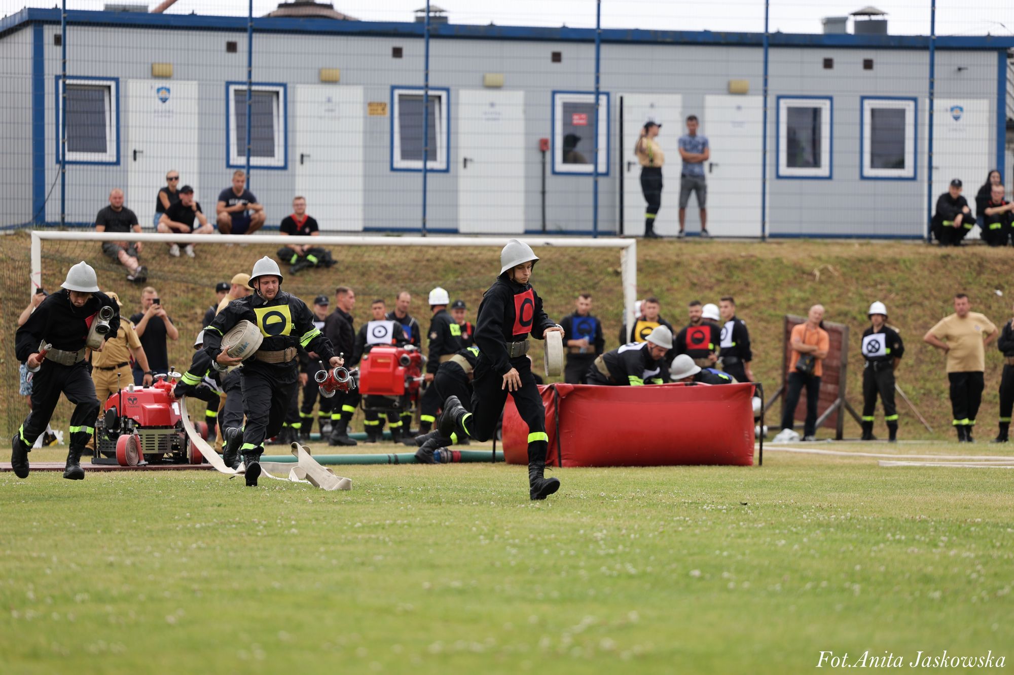 Trzech strażaków w hełmach i czarnych mundurach z kolorowymi oznaczeniami biegnie po trawiastym boisku z wężami strażackimi, w tle ludzie i kontenery.