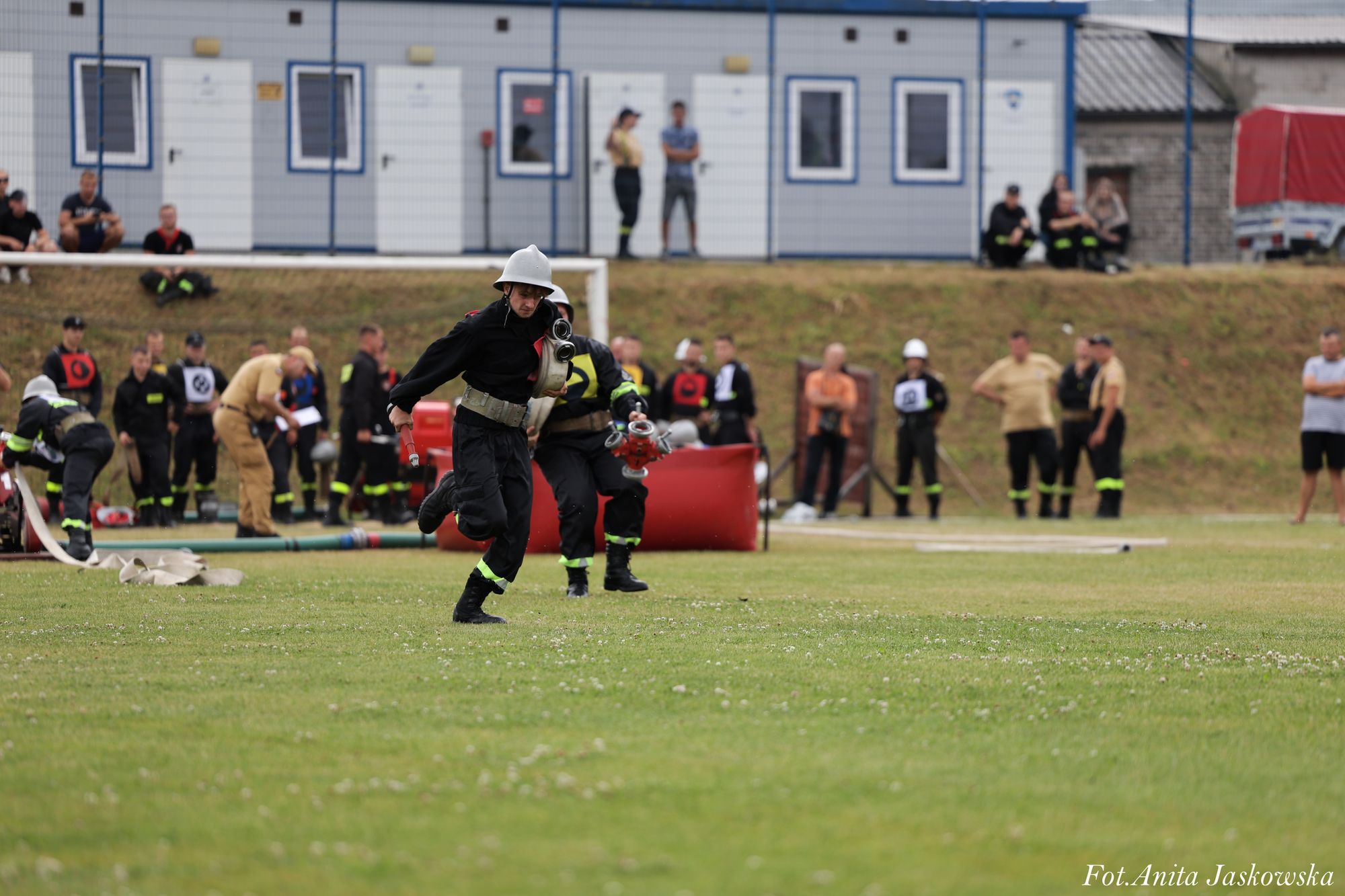Dwóch strażaków w ciemnych mundurach i białych hełmach biegnie po trawie z wężem strażackim, w tle grupa ludzi obserwujących wydarzenie przy budynkach i ogrodzeniu.