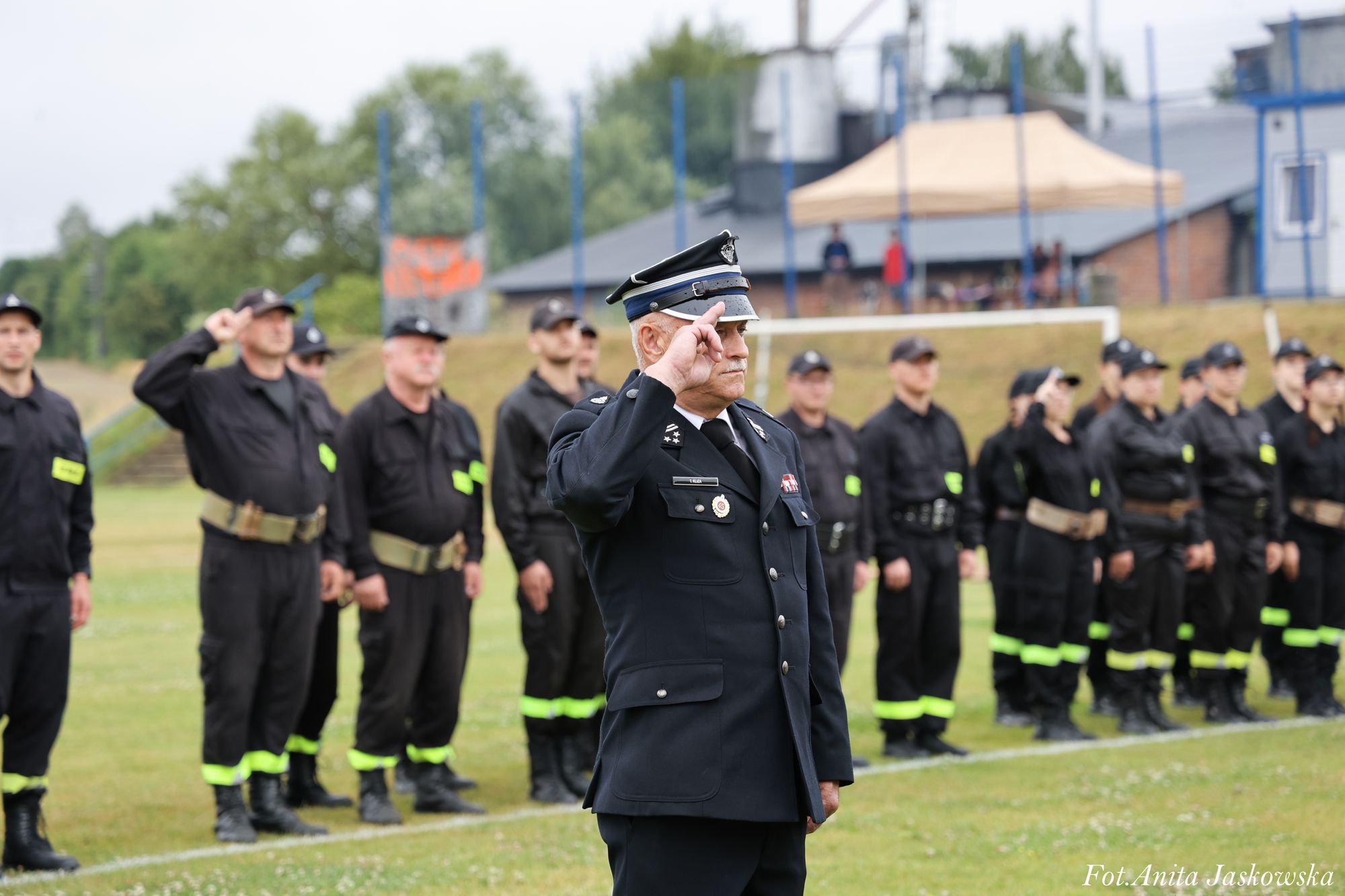 Mężczyzna w granatowym mundurze z czapką w geście salutowania, w tle grupa osób w czarnych uniformach z żółtymi opaskami, trawa i budynki.
