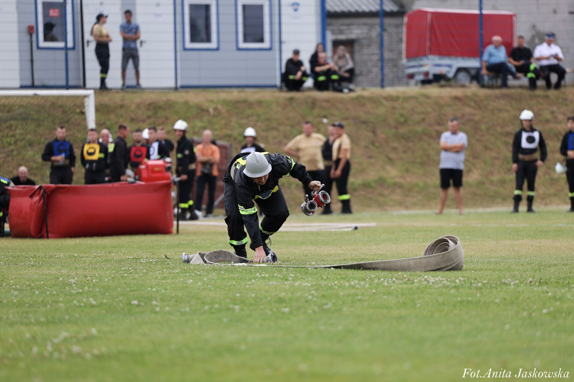 Strażak w kasku i ubraniu ochronnym rozwija zwinięty wąż strażacki na zielonej trawie, w tle grupa osób i sprzęt strażacki.
