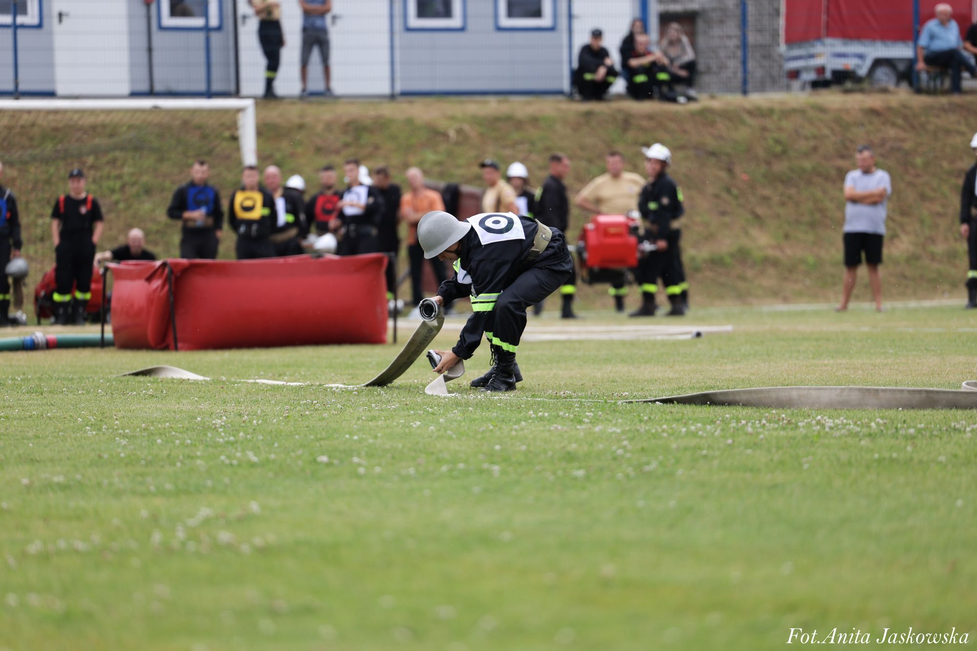 Strażak w czarnym mundurze i białym hełmie z numerem 0 na plecach rozkłada na trawie zwinięty wąż strażacki, w tle grupa osób i czerwony sprzęt.