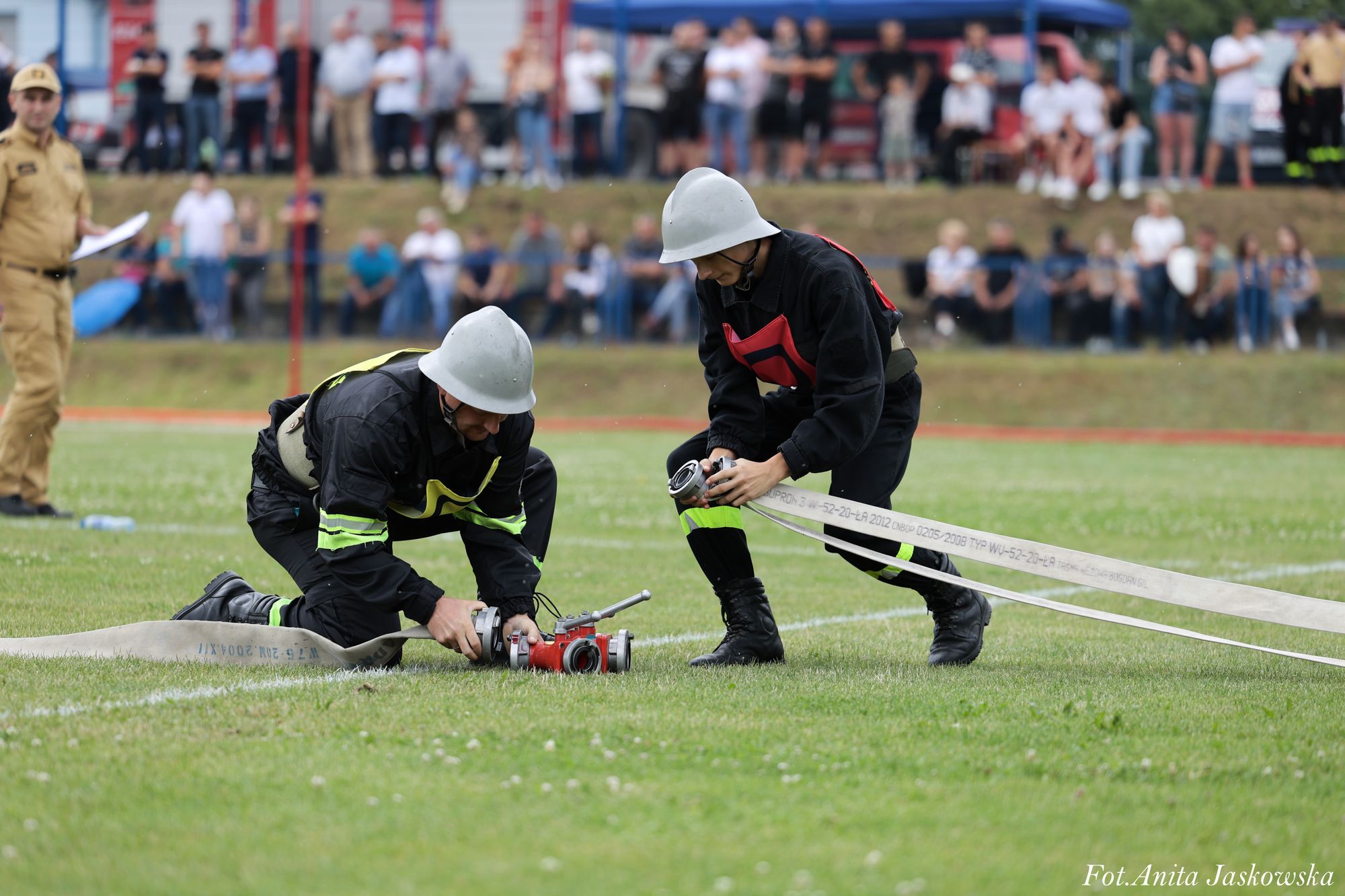 Dwóch strażaków w czarnych mundurach i białych hełmach montuje sprzęt pożarniczy na zielonej trawie, w tle rozmyta grupa ludzi i mężczyzna w beżowym stroju z dokumentami.