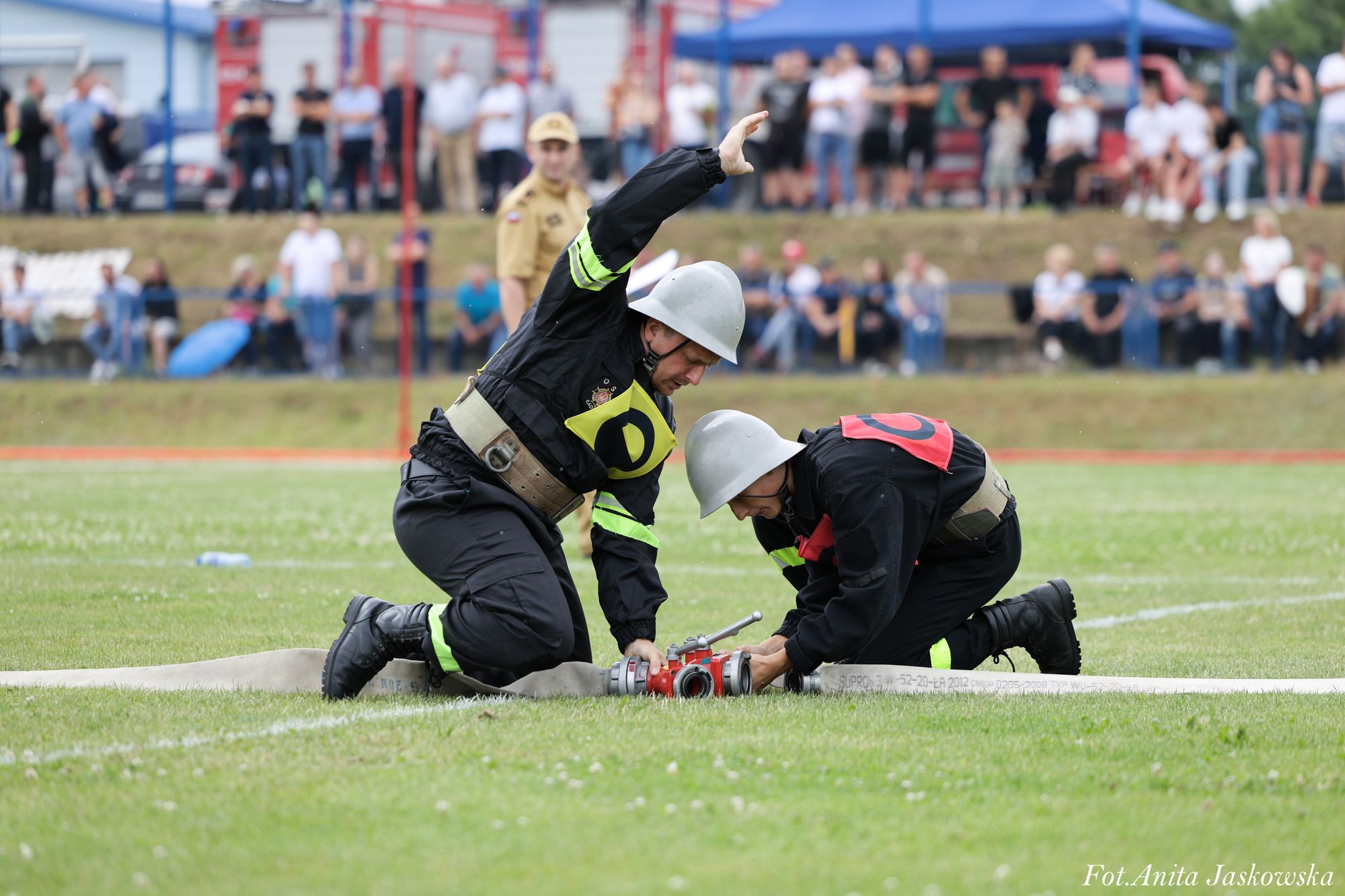Dwóch strażaków w czarnych mundurach i hełmach klęczy na trawie, łącząc węże strażackie, w tle rozmyta grupa ludzi.