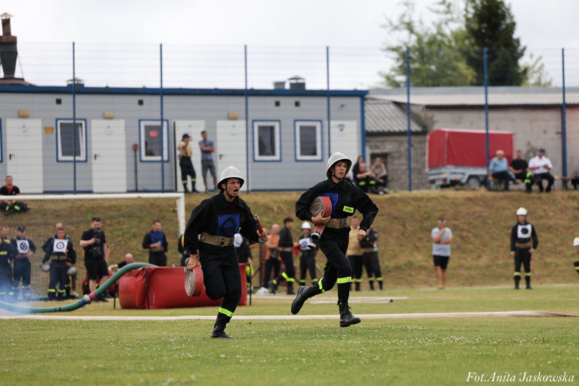 Dwóch mężczyzn w czarnych mundurach strażackich z białymi hełmami biegnie po zielonym boisku, każdy trzyma zwinięty wąż strażacki, w tle ludzie i budynki.