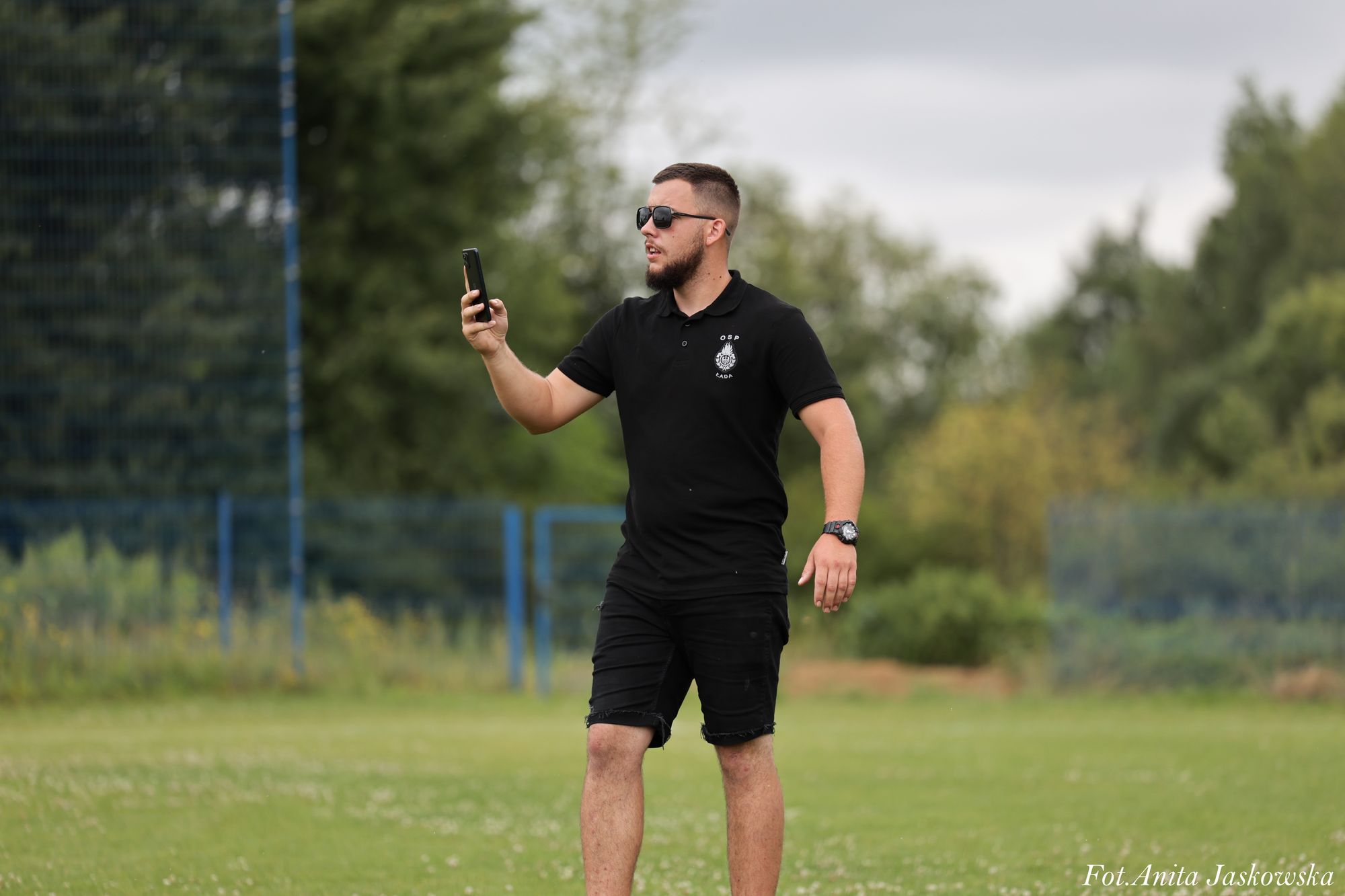 Mężczyzna w czarnych okularach, czarnej koszulce i czarnych szortach stoi na zielonej trawie, trzymając telefon na tle rozmytych drzew i nieba.