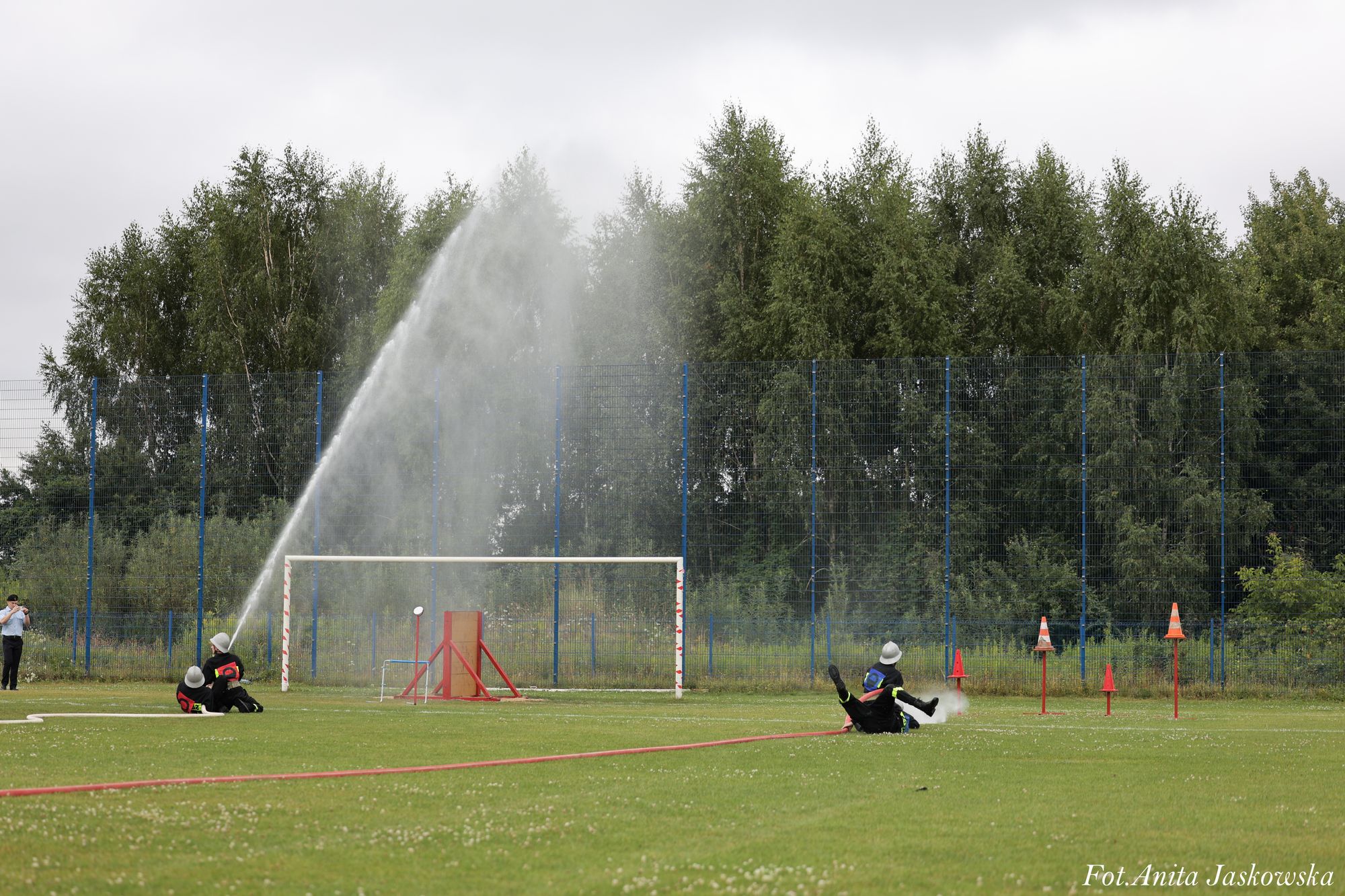 Trzech strażaków w czarnych mundurach i hełmach na zielonym boisku, jeden strzela strumieniem wody z węża, w tle bramka i metalowe płotki, z tyłu drzewa i pochmurne niebo.