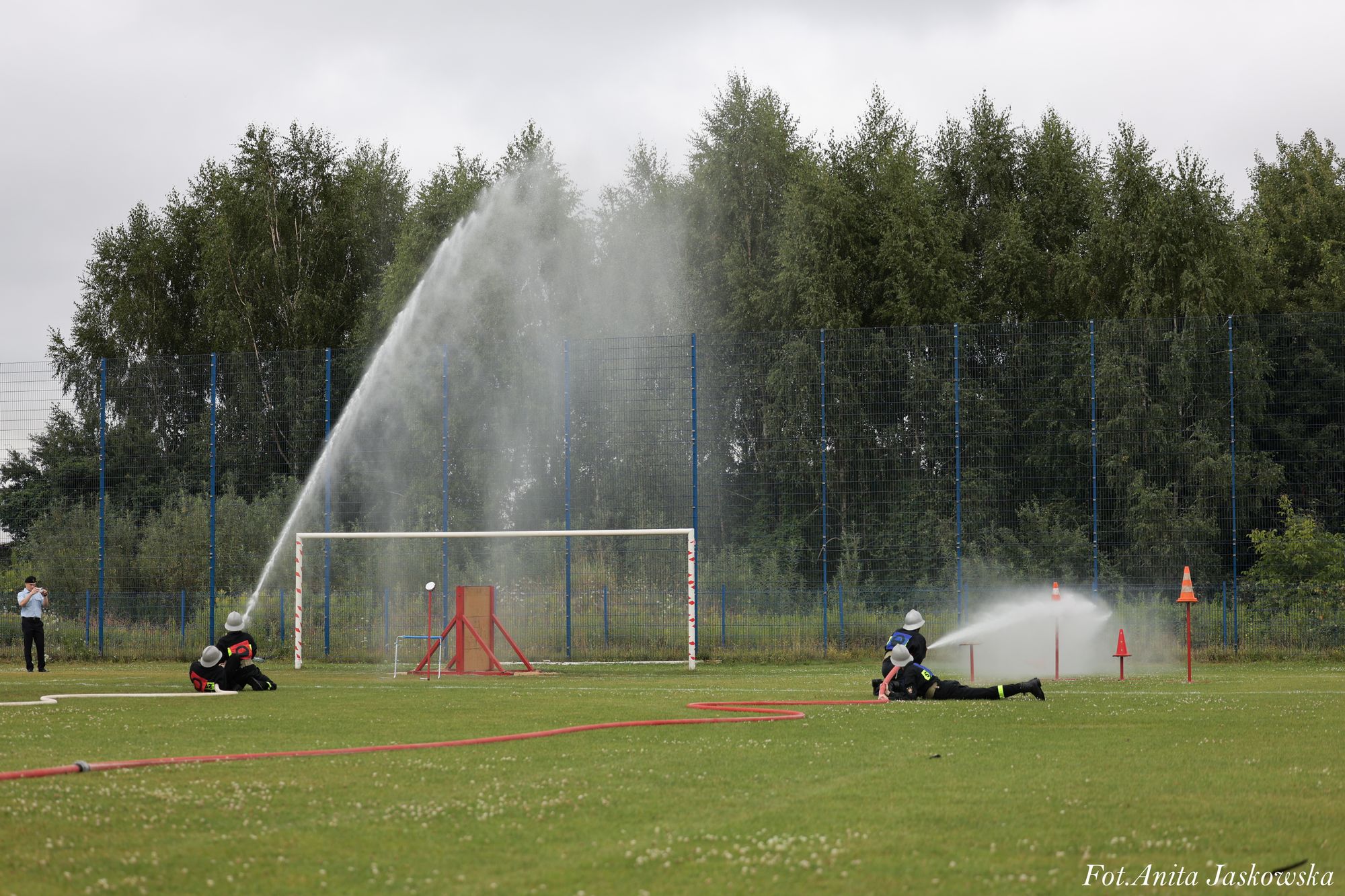 Drużyna strażaków w hełmach na zielonym boisku, strumienie wody z węży gaśniczych skierowane na tarcze, w tle ogrodzenie i drzewa, pochmurne niebo.
