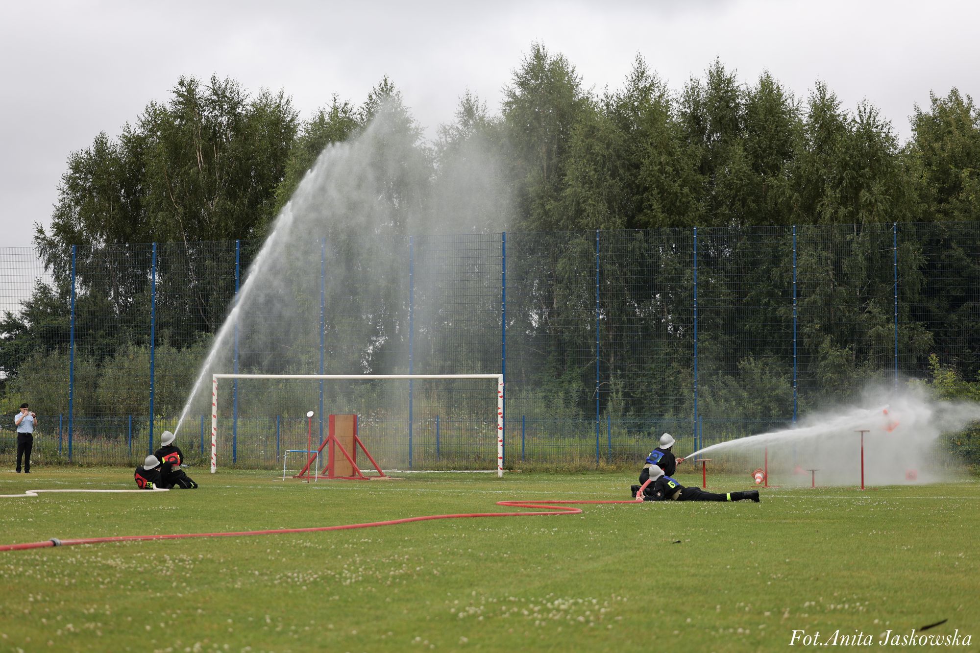 Dwie pary strażaków ćwiczące gaszenie wodą na zielonym boisku, za nimi bramka i wysoka siatka, w tle drzewa i pochmurne niebo.