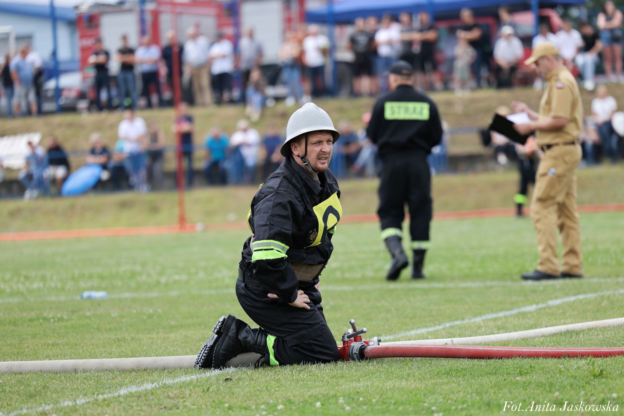 Strażak w czarnym ubraniu ochronnym i hełmie klęczy na zielonej trawie obok czerwonego węża strażackiego, w tle rozmyci ludzie i sędzia w beżowym mundurze.