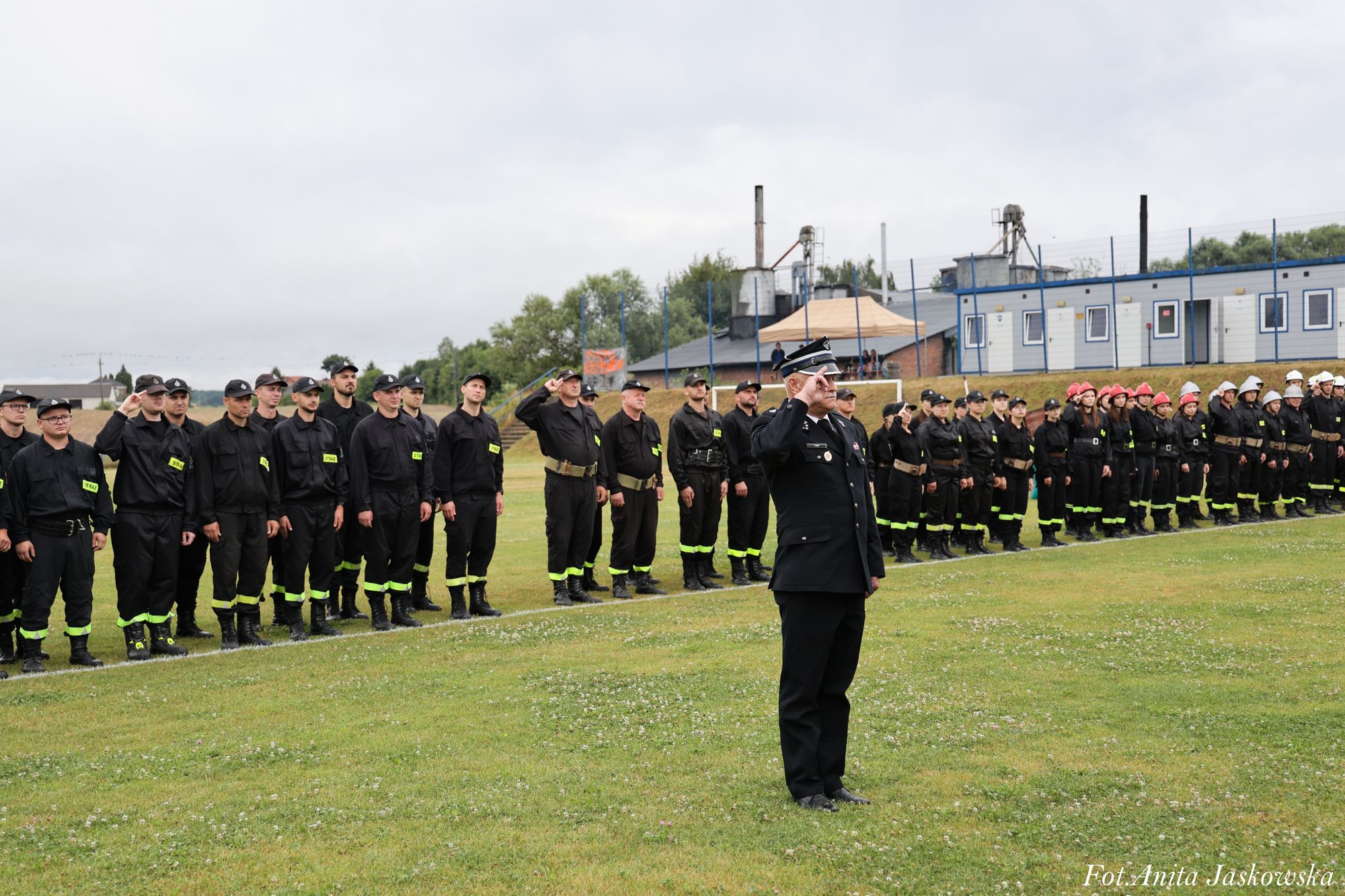 Grupa osób w czarnych mundurach z odblaskowymi paskami, jedne z nich w kaskach strażackich, stoją na trawie w rzędach, mężczyzna w mundurze oficerskim salutuje.