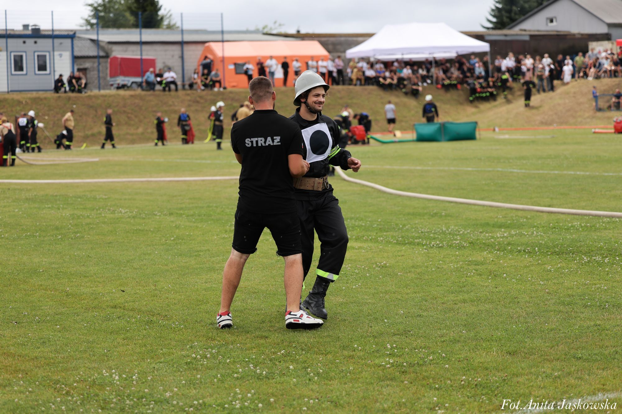 Dwóch mężczyzn na zielonym boisku, jeden w czarnych spodniach i białych butach z napisem "STRAŻ" na koszulce, drugi w czarnym ubraniu strażaka i hełmie.