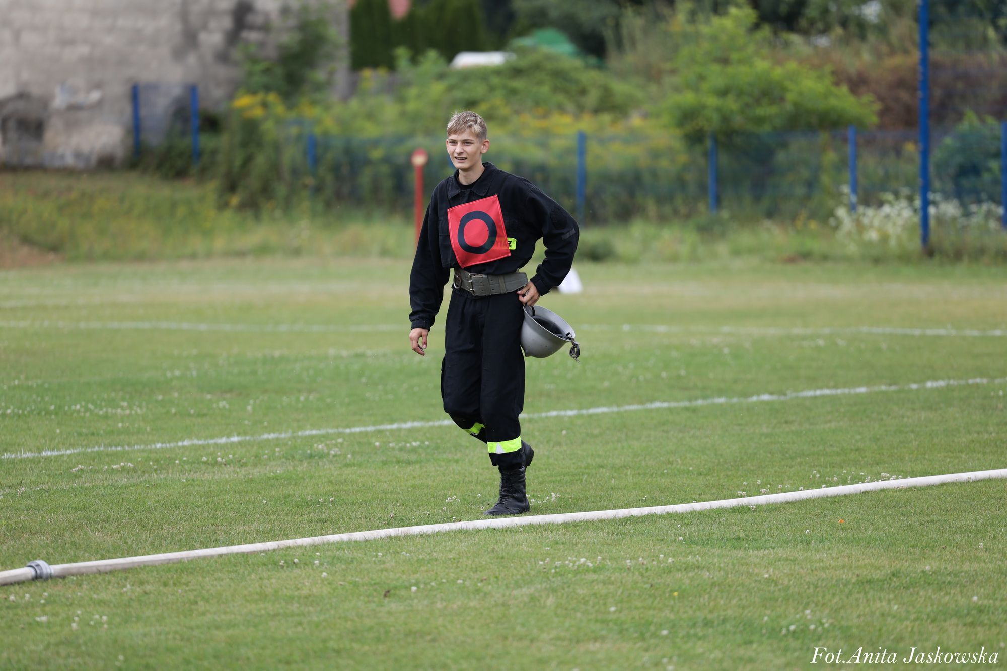 Młody mężczyzna w czarnym ubraniu ochronnym z odblaskowymi paskami, trzymający szary hełm, stoi na trawiastym boisku obok białej rury.